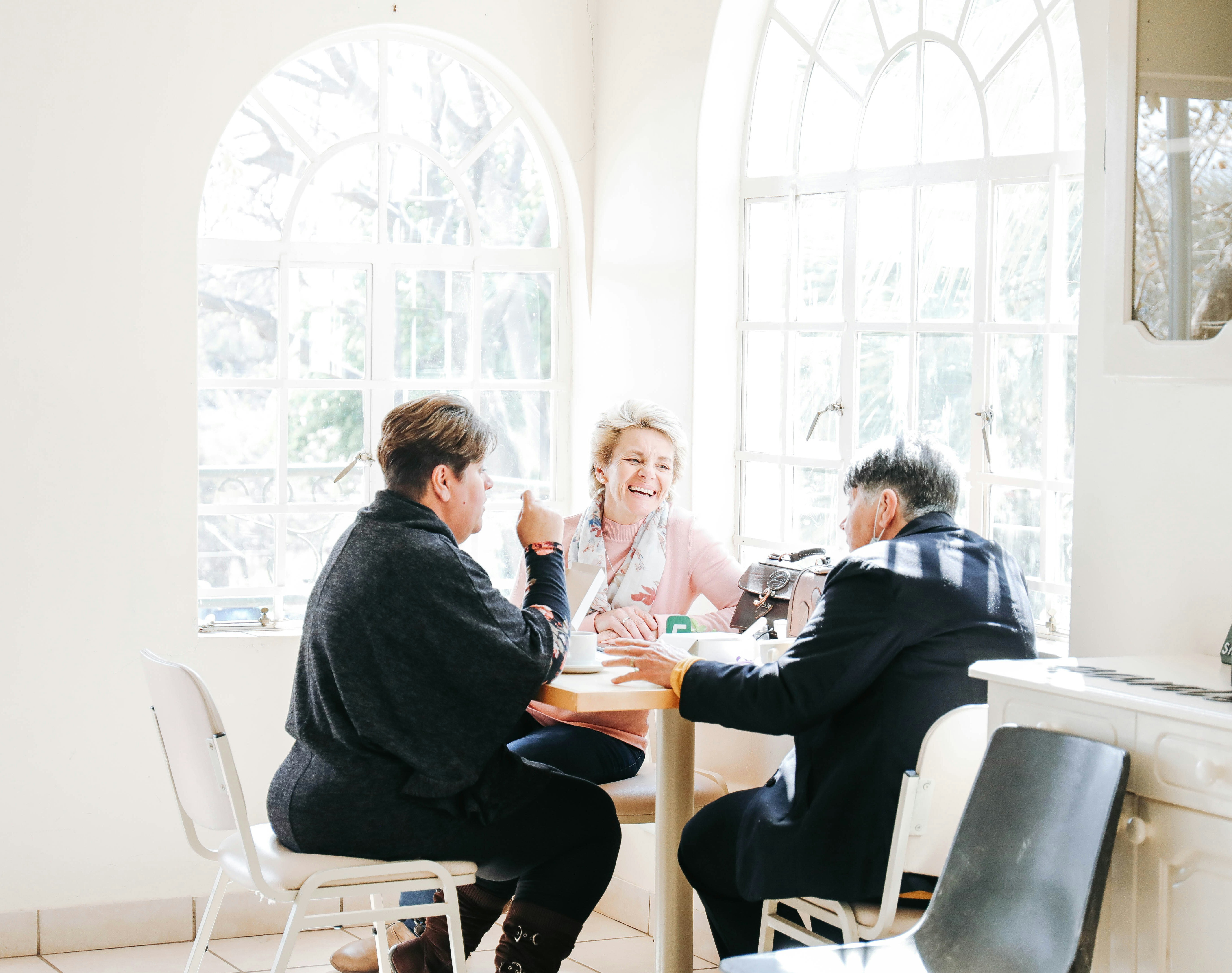 Three older adults engaged in a friendly and joyful conversation at a sunlit café, sitting around a table with warm smiles and relaxed body language, creating a welcoming and supportive atmosphere. Three older adults engaged in a friendly and joyful conversation at a sunlit café, sitting around a table with warm smiles and relaxed body language, creating a welcoming and supportive atmosphere.