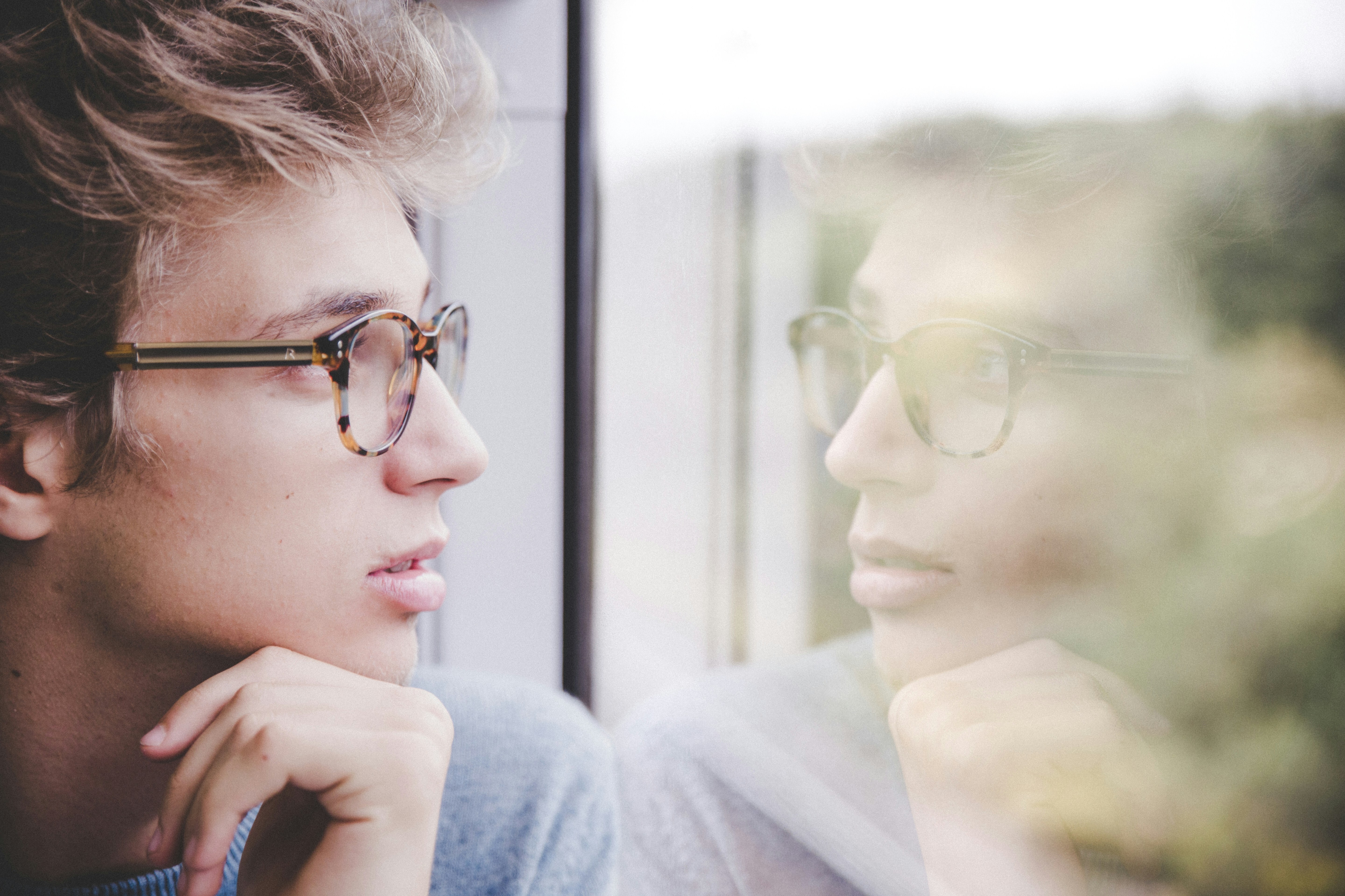 Young man wearing glasses, gazing thoughtfully out of a window, with his reflection visible on the glass, creating a contemplative and introspective mood. Young man wearing glasses, gazing thoughtfully out of a window, with his reflection visible on the glass, creating a contemplative and introspective mood.