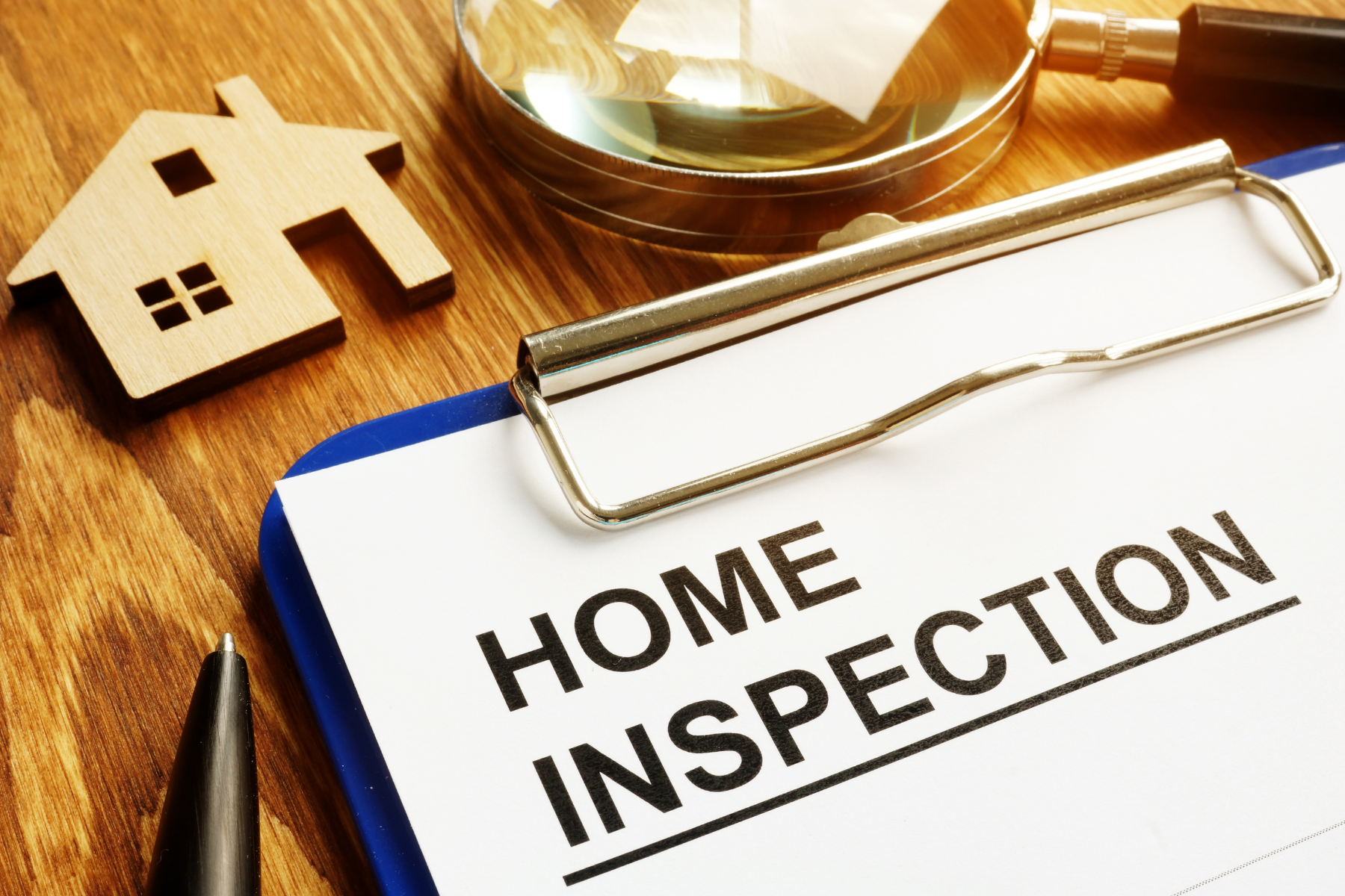 The image features a clipboard labeled "HOME INSPECTION" alongside a wooden house cutout, magnifying glass, and pen—representing the critical step of evaluating a property before buying or selling. The image features a clipboard labeled "HOME INSPECTION" alongside a wooden house cutout, magnifying glass, and pen—representing the critical step of evaluating a property before buying or selling.