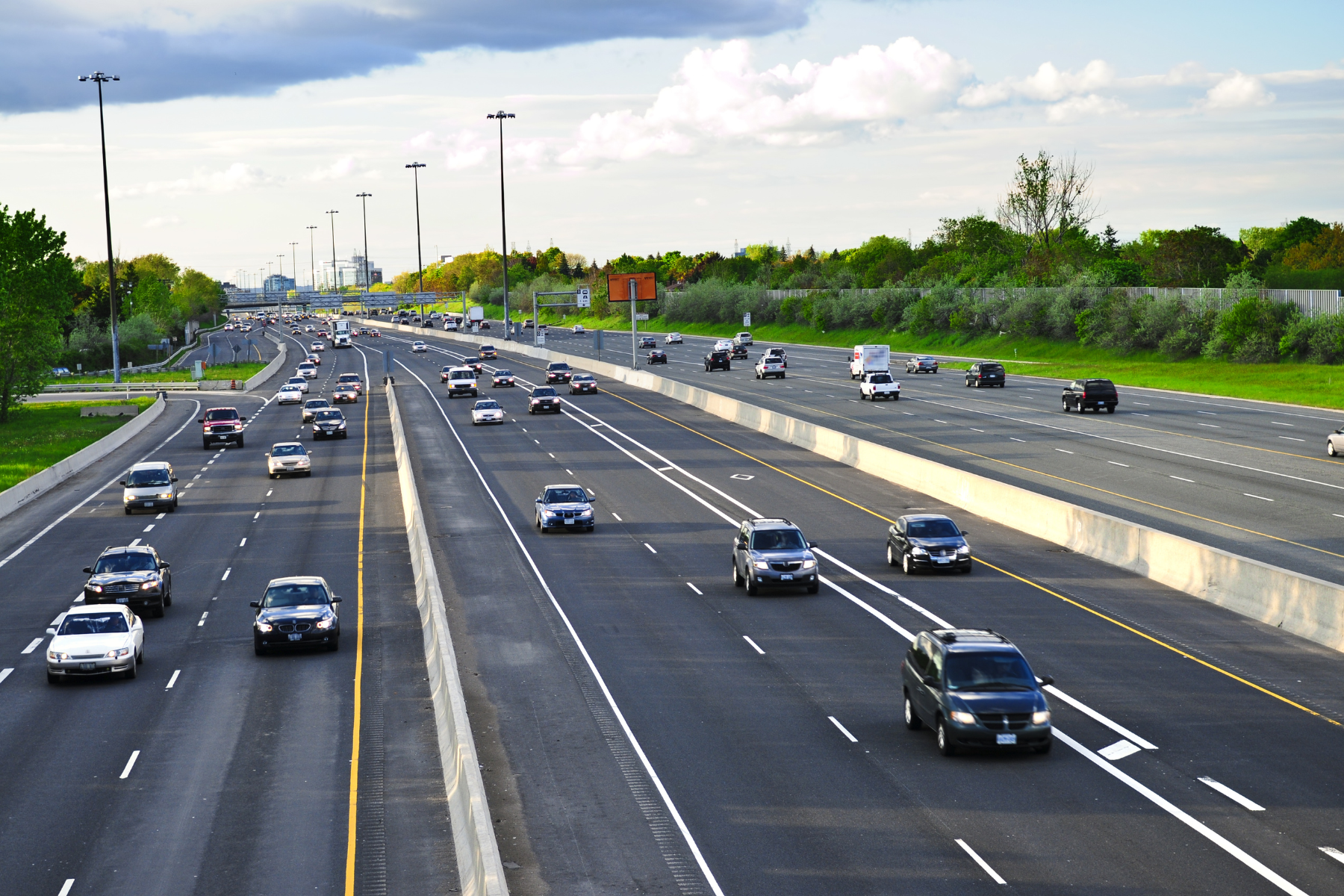 The image shows a busy multi-lane highway with cars traveling in both directions, bordered by greenery and trees, under a partly cloudy sky. The image shows a busy multi-lane highway with cars traveling in both directions, bordered by greenery and trees, under a partly cloudy sky.