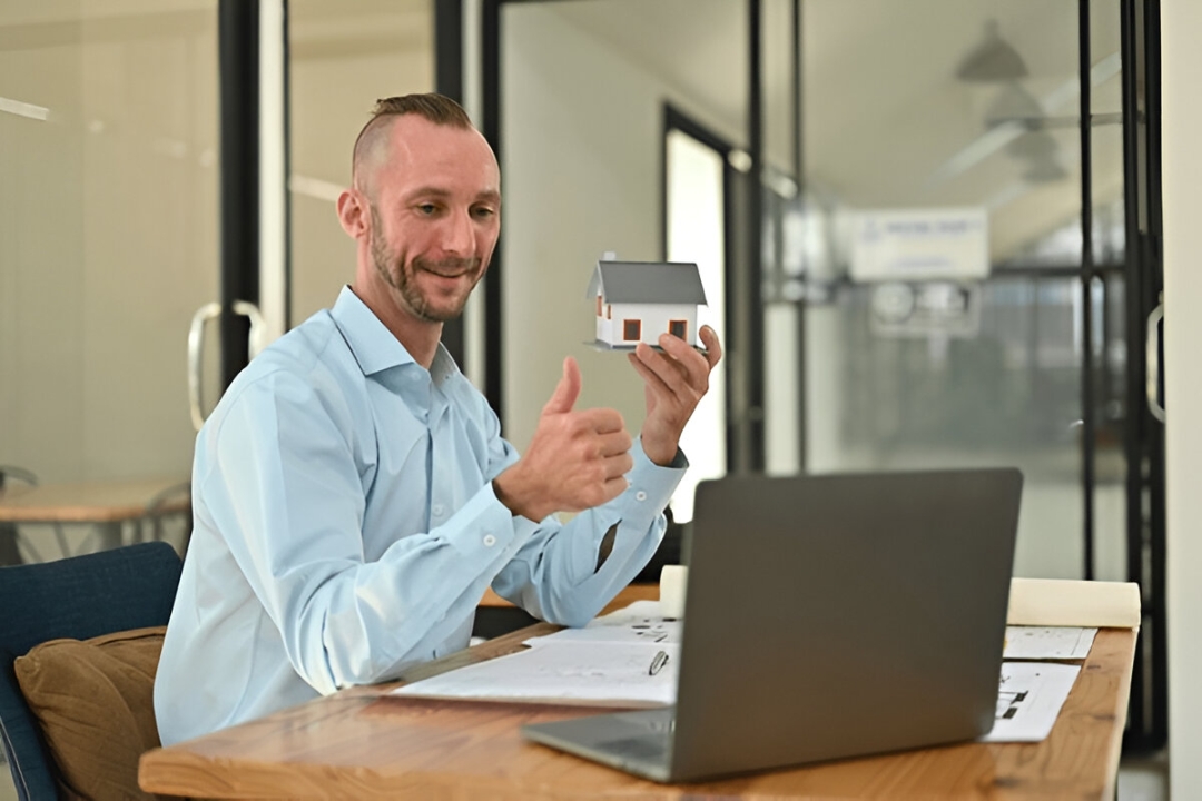 A realtor on a video call gives a thumbs-up while holding a model house, demonstrating video marketing for property investors from AMPLIFIREI.