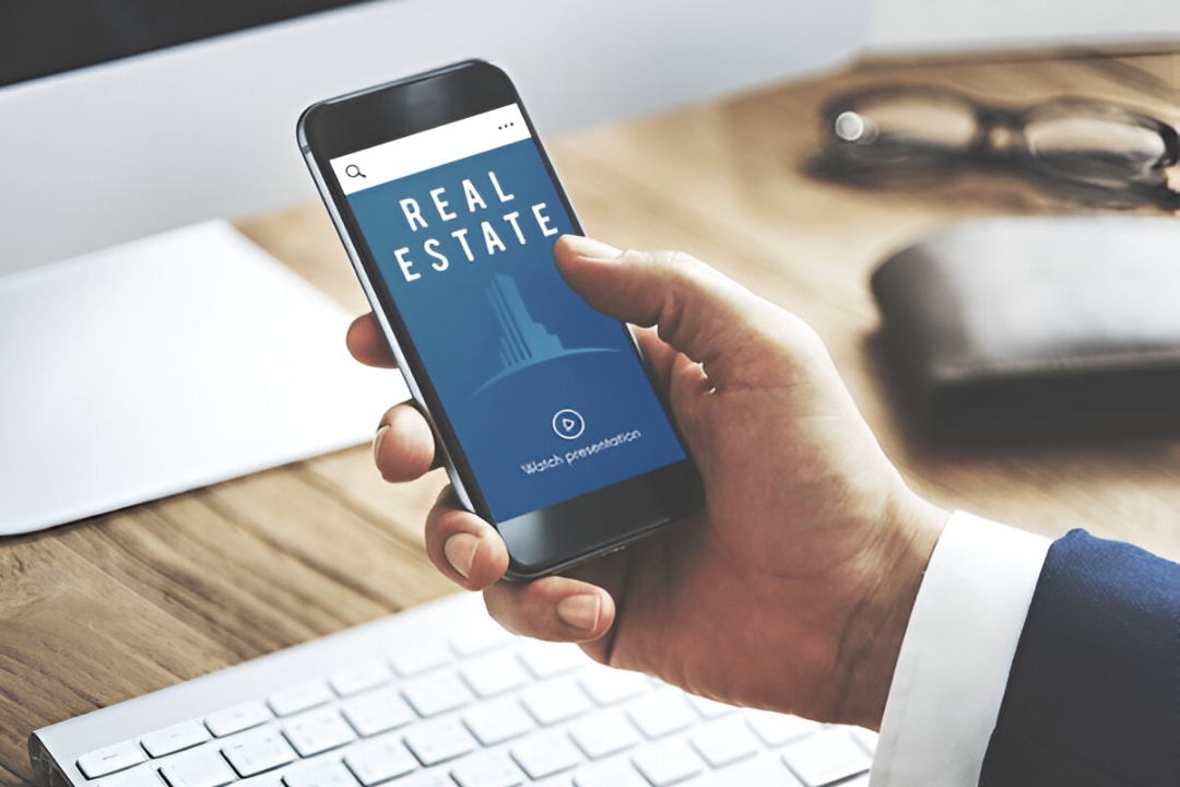 A close-up of a man's hand holding a smartphone displaying a "REAL ESTATE" app, showcasing video marketing for property investors from AMPLIFIREI.