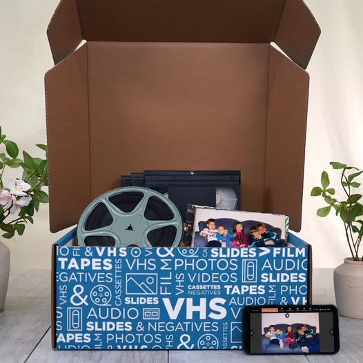 Open digitization box filled with old media formats including a film reel, VHS tapes, printed photos, and a tablet displaying a scanned image, with decorative plants in the background. Open digitization box filled with old media formats including a film reel, VHS tapes, printed photos, and a tablet displaying a scanned image, with decorative plants in the background.