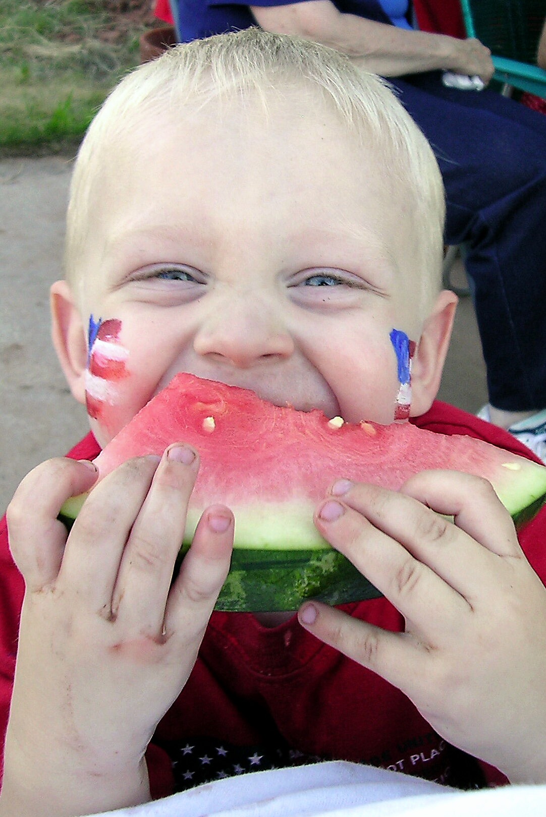 A young child with blonde hair and face paint in red, white, and blue, happily eating a large slice of watermelon with both hands. The child is wearing a red shirt and smiling with squinted eyes. A young child with blonde hair and face paint in red, white, and blue, happily eating a large slice of watermelon with both hands. The child is wearing a red shirt and smiling with squinted eyes.