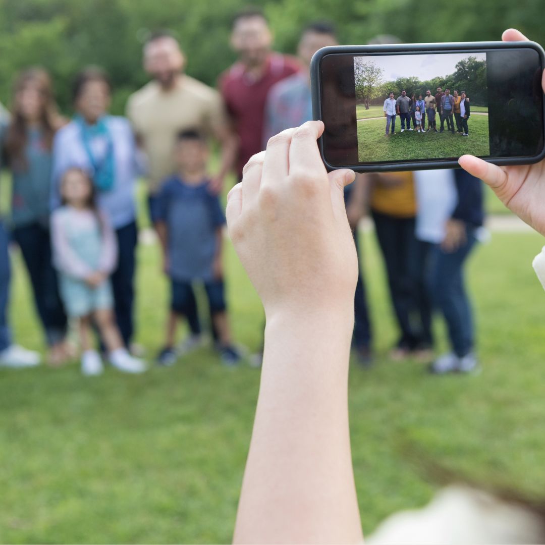 Close-up of a person’s hand holding a smartphone, taking a photo of a multigenerational family group standing together on grass in a park. Close-up of a person’s hand holding a smartphone, taking a photo of a multigenerational family group standing together on grass in a park.