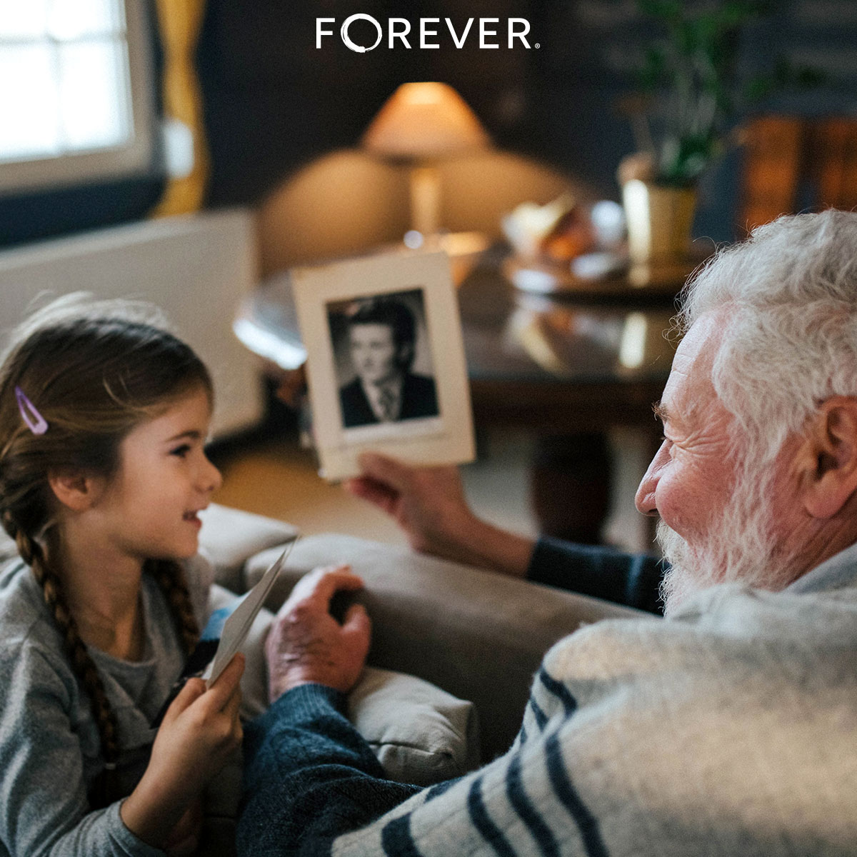 An elderly man smiles while showing a young girl a black-and-white portrait photograph, creating a moment of storytelling and connection across generations. The FOREVER® logo is visible at the top.