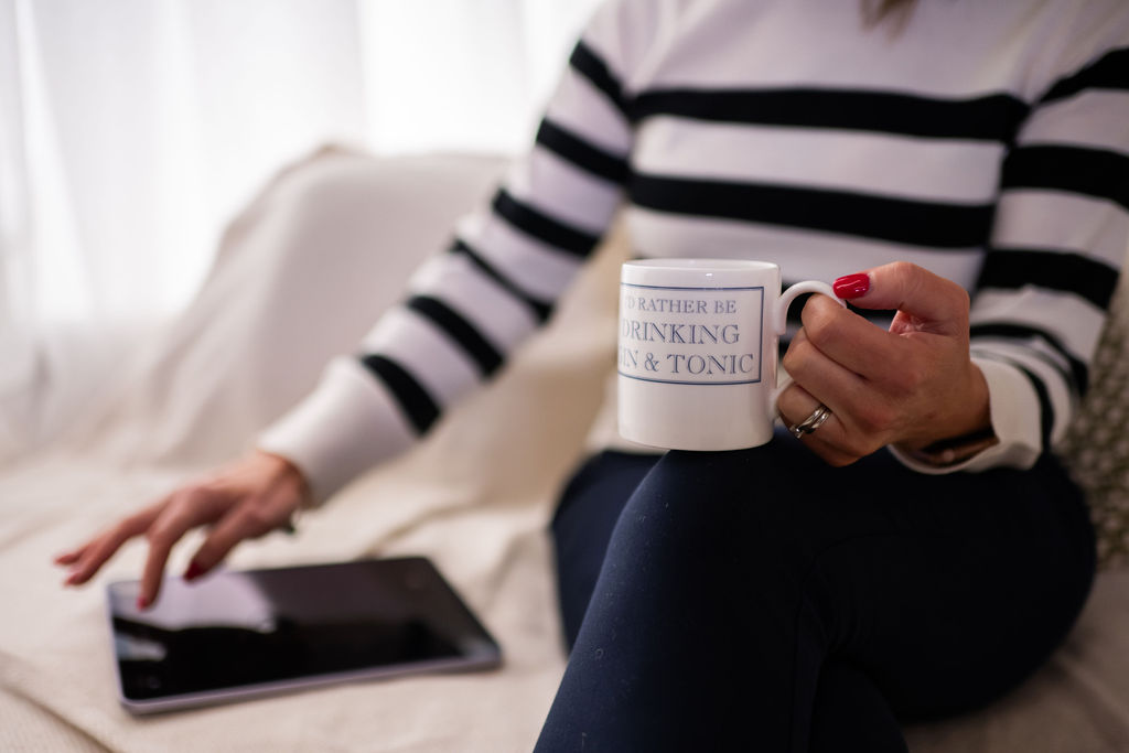 Woman sitting with a cup that says "I'd rather be drinking gin & tonic," holding a tablet on her lap