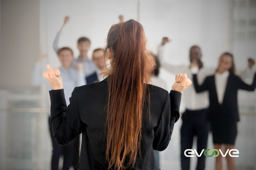 "A confident woman in a black blazer stands with her back to the camera, raising her fists in victory, while a group of diverse professionals in the background cheers in support. The Evoove logo is placed in the bottom right corner, symbolizing leadership, empowerment, and team success. "A confident woman in a black blazer stands with her back to the camera, raising her fists in victory, while a group of diverse professionals in the background cheers in support. The Evoove logo is placed in the bottom right corner, symbolizing leadership, empowerment, and team success.