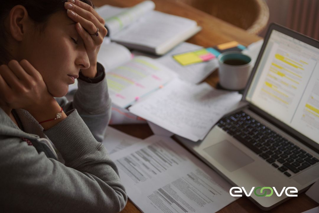 A stressed professional sits at a cluttered desk with open books, documents, and a laptop, overwhelmed by workload and deadlines, with the Evoove logo representing staffing solutions for balance and efficiency.