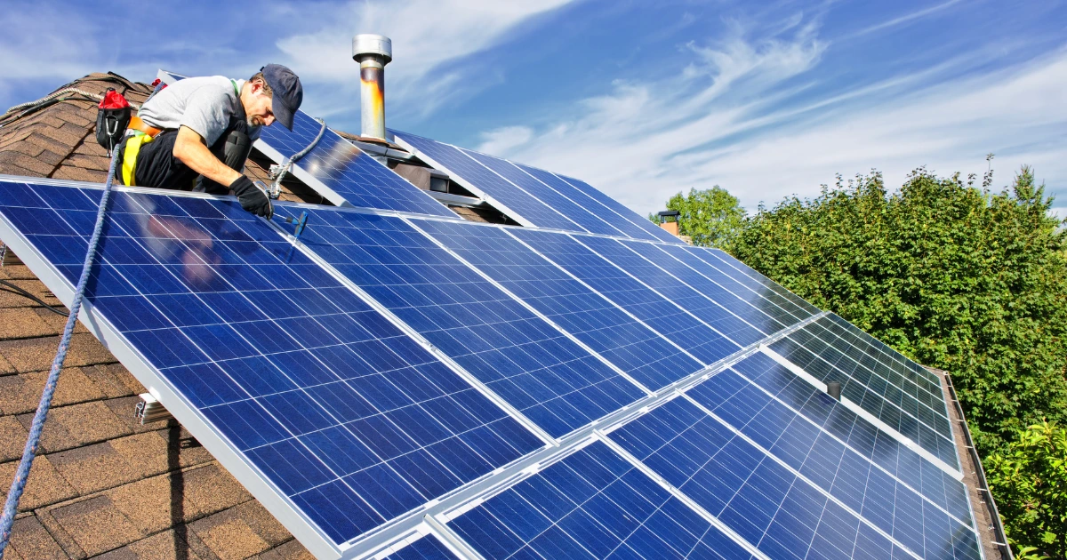 A man installing a solar panel on a rooftop, focused on his work under a clear blue sky.  