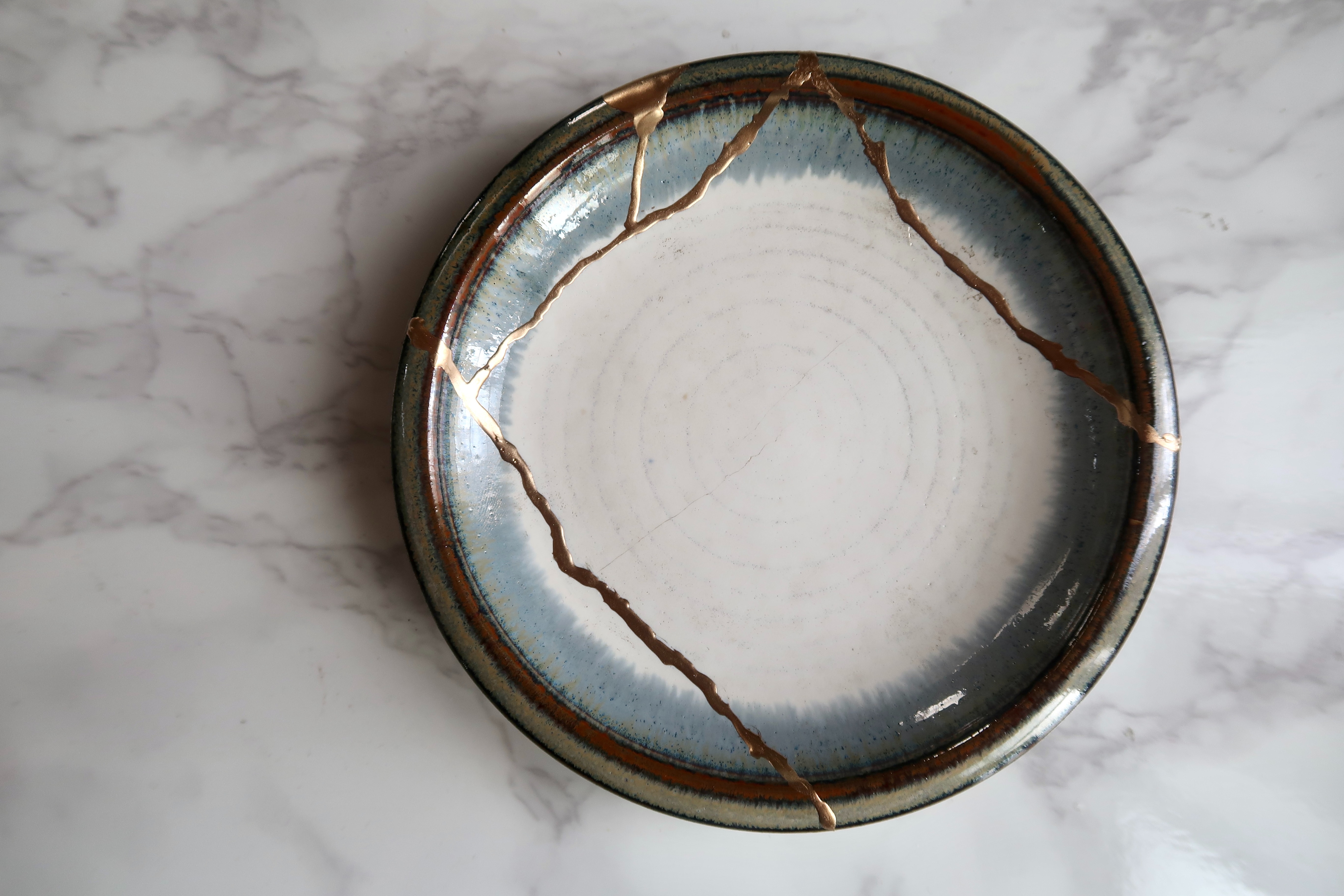 A beige-colored plate with a ring of pale blue, edged in gold, with beautiful gold-filled cracks. Image credit: Riho Kitagawa on Unsplash