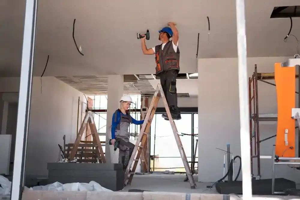 Two construction workers installing ceiling panels on ladders inside a building under renovation.