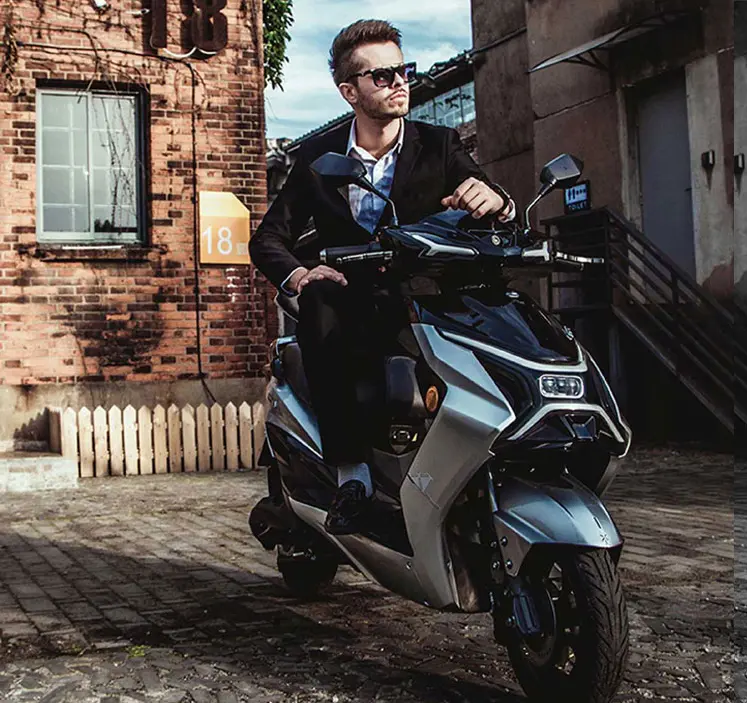 Moški v elegantni črni obleki sedi na električnem skuterju VeloVoltage HyperX, parkiranem na tlakovani ulici ob stari opečnati stavbi, z odločnim pogledom usmerjenim naprej.
