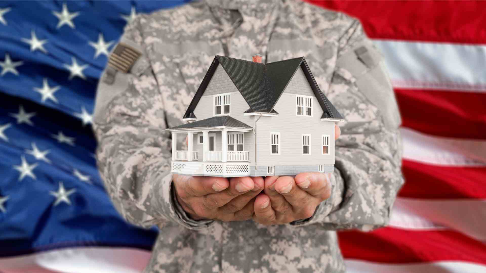 A soldier holds up a small house in front of an American Flag. The home and flag represent the process of obtaining home financing through a VA loan and closing on a new home purchase.