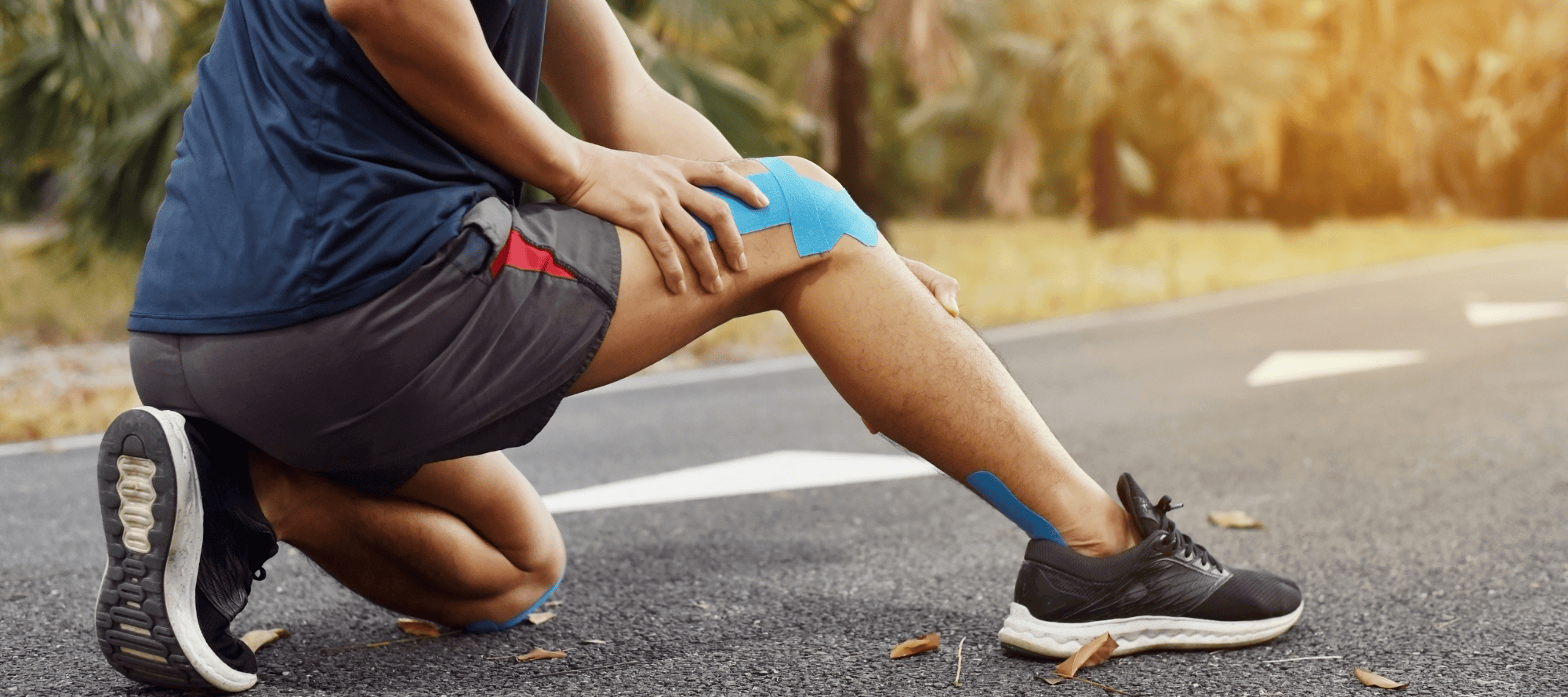 A runner on a road is crouched down, gripping their left knee which is supported by blue kinesiology tape, indicating a possible injury or strain. The person is wearing black shorts, a navy blue shirt, and black athletic shoes, and appears to be in discomfort, highlighting the focus of the blog article on common knee issues post-surgery and the importance of proper knee care.