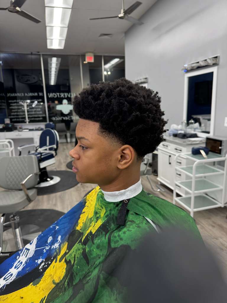 Wide shot of the barbershop interior with warm wood counters and mirrored stations, three barbers working, natural morning light, photorealistic editorial emphasizing community and craft.