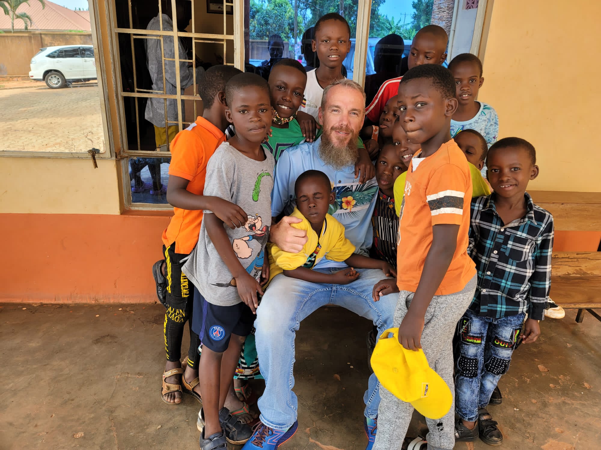 Group of children gathered around an adult outdoors, smiling and posing for a photo in front of a building.