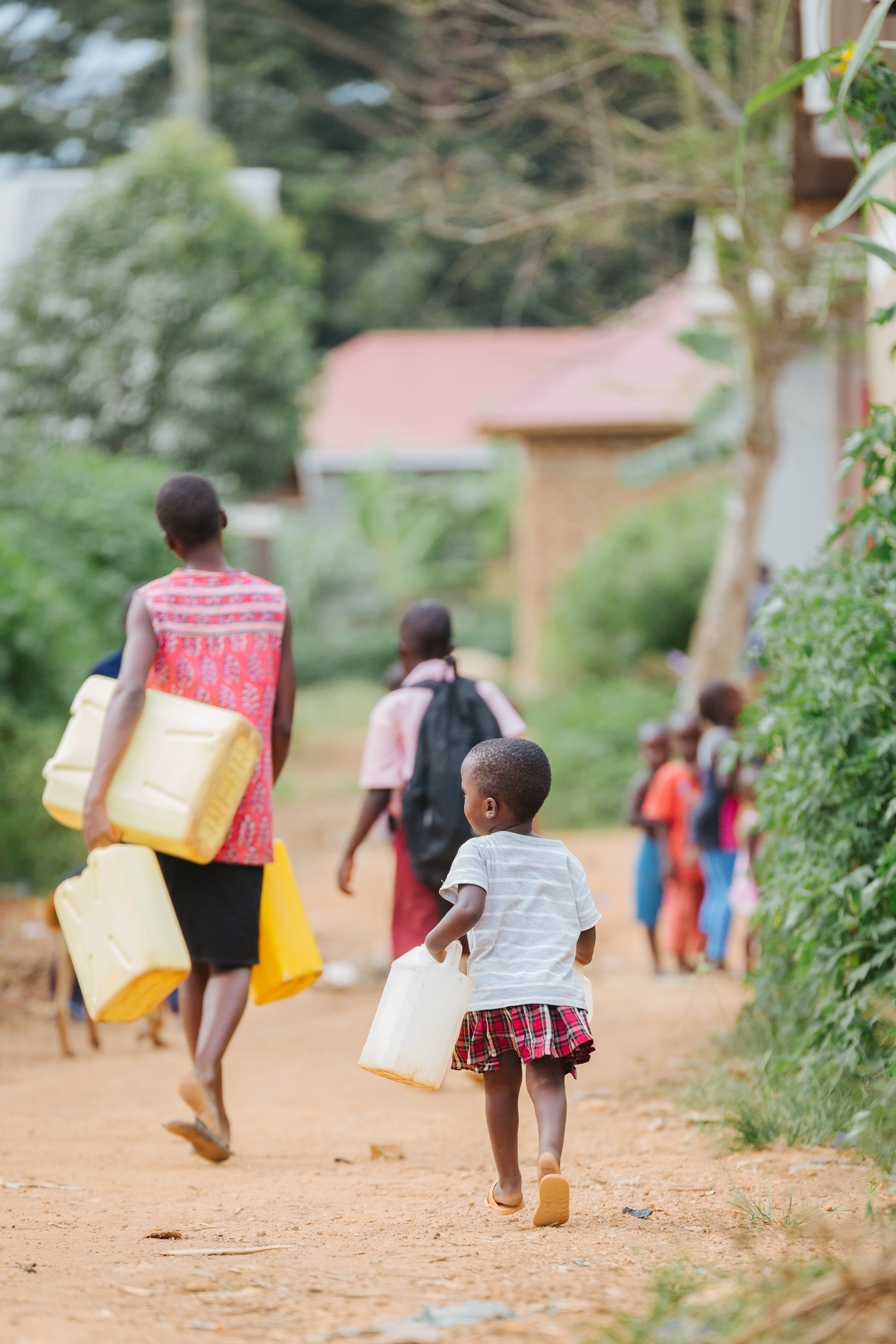 Small child carrying a white water jug walking behind an adult carrying yellow jerrycans on a dirt road in a Ugandan village.