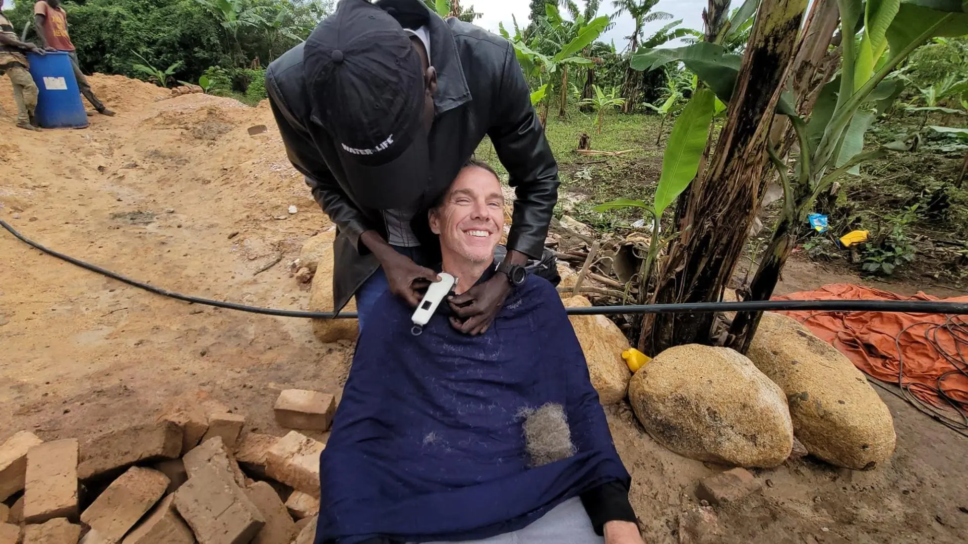 Smiling man receiving a haircut outdoors in a rural village, with construction bricks and banana trees in the background.