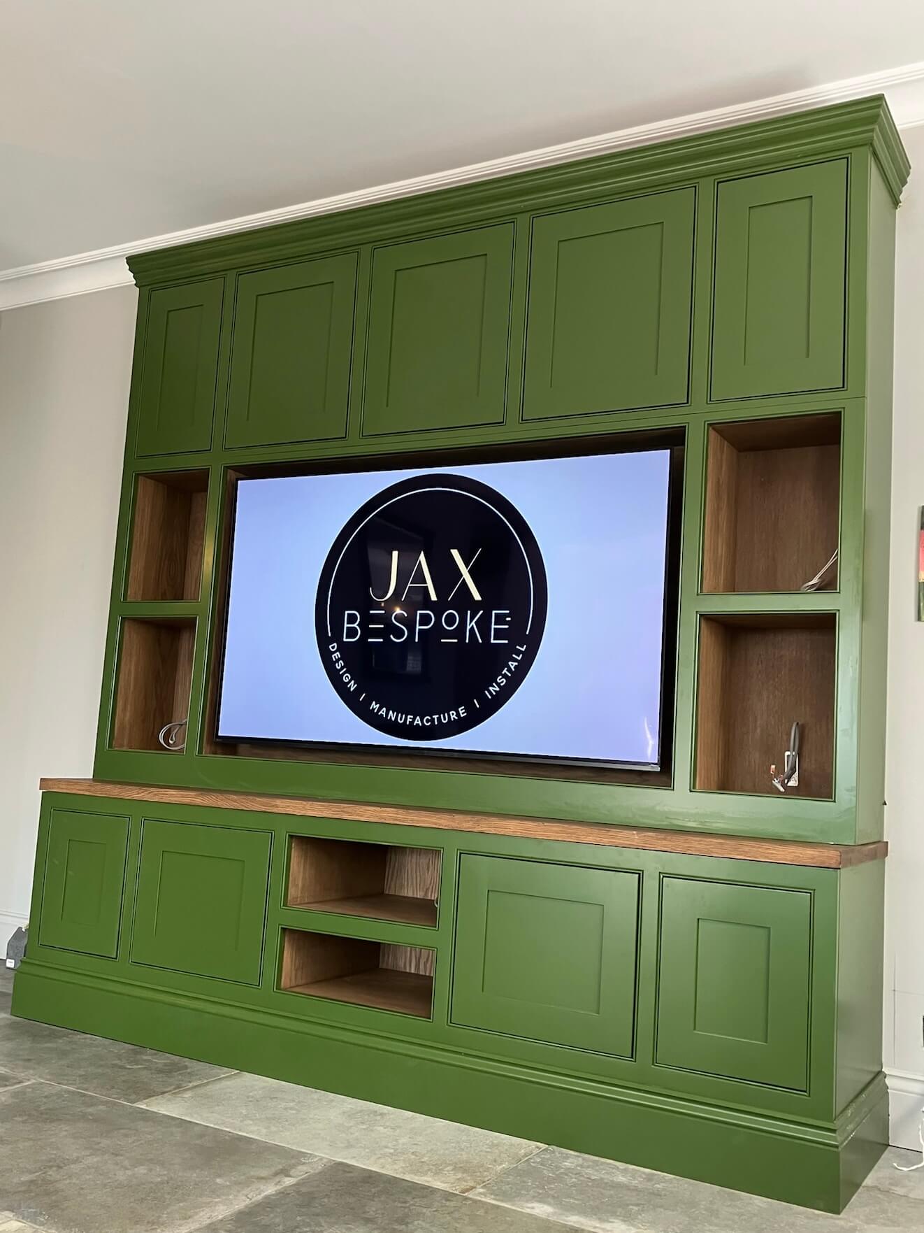 Harrogate Custom built media wall unit in sage green with oak accents featuring a TV flanked by decorative alcoves with oak shelving. The design includes paneled cabinet doors above and below the TV area, with additional open shelving spaces in oak finish Harrogate Custom built media wall unit in sage green with oak accents featuring a TV flanked by decorative alcoves with oak shelving. The design includes paneled cabinet doors above and below the TV area, with additional open shelving spaces in oak finish
