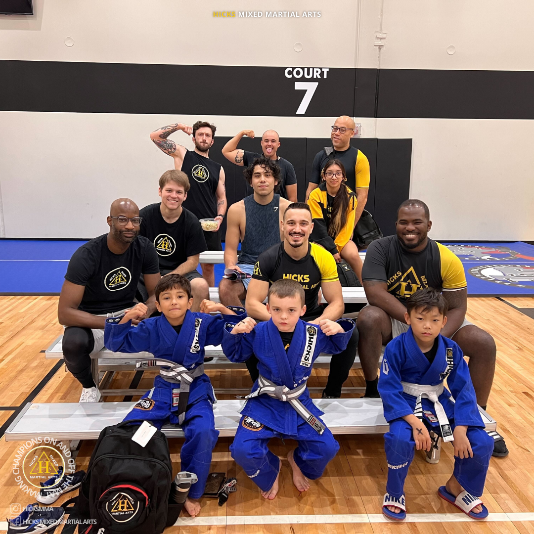 Adults and kids at a martial arts tournament representing Hicks MMA, showcasing the community, progression, and teamwork in BJJ and martial arts. Adults and kids at a martial arts tournament representing Hicks MMA, showcasing the community, progression, and teamwork in BJJ and martial arts.