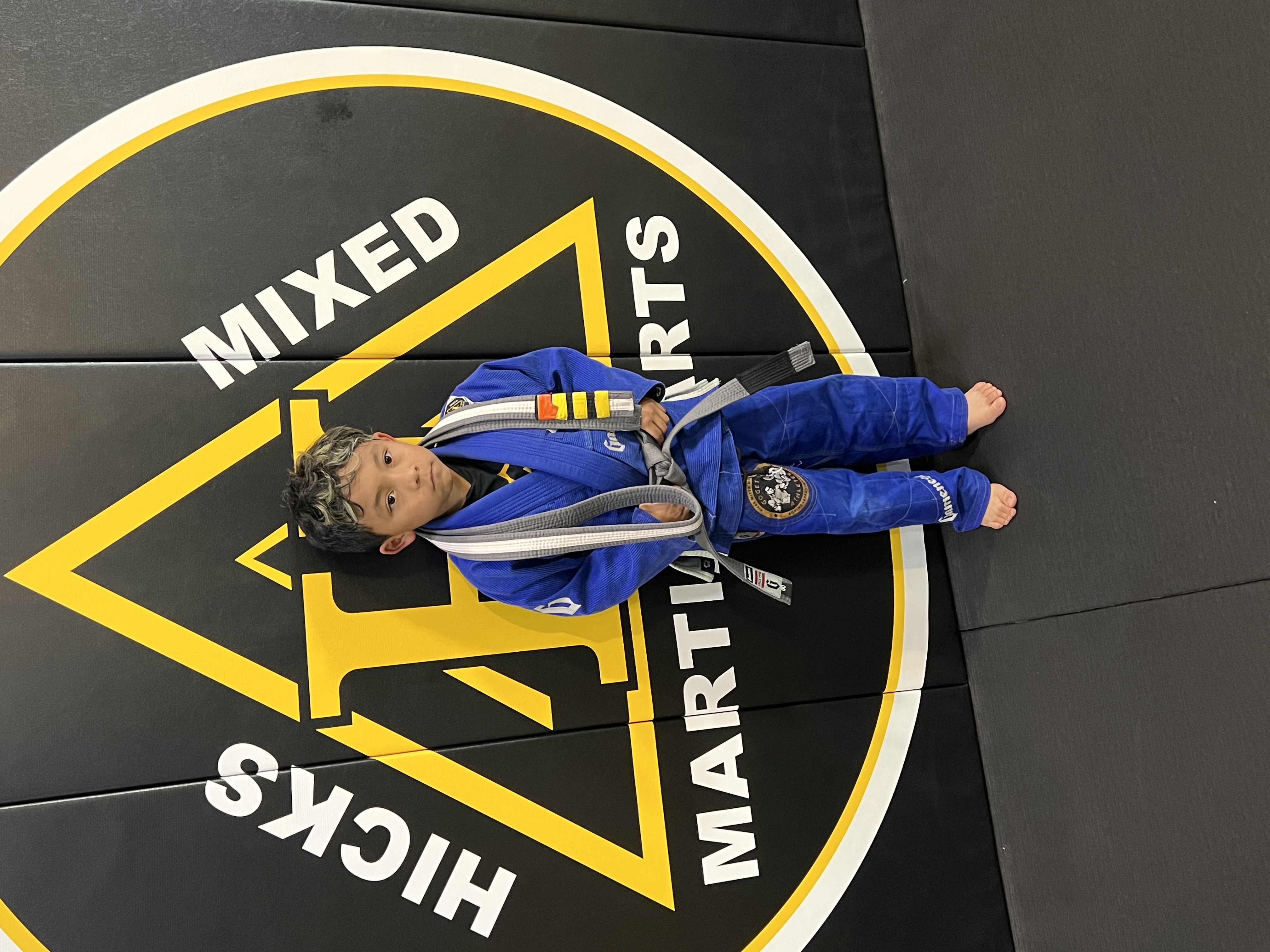 Child being promoted from grey-white belt to grey belt in Brazilian Jiu-Jitsu at Hicks MMA, showcasing growth, discipline, and progression in martial arts training. Child being promoted from grey-white belt to grey belt in Brazilian Jiu-Jitsu at Hicks MMA, showcasing growth, discipline, and progression in martial arts training.