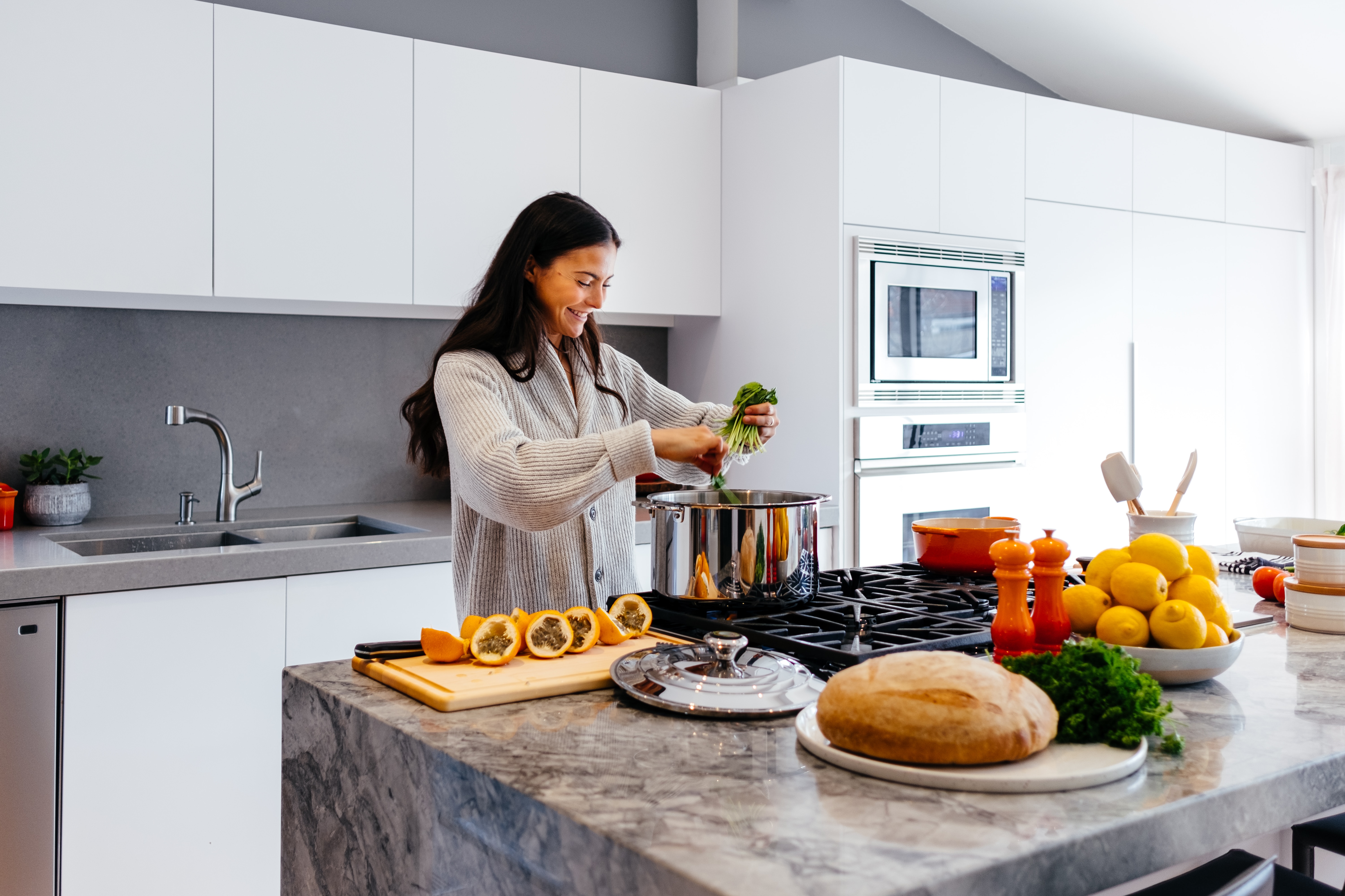 A woman with long dark hair, wearing a white sweater, cooks in a kitchen.  The counter has sliced squash, a bowl of lemons, and a loaf of bread, and she's putting green vegetables into a large pot.