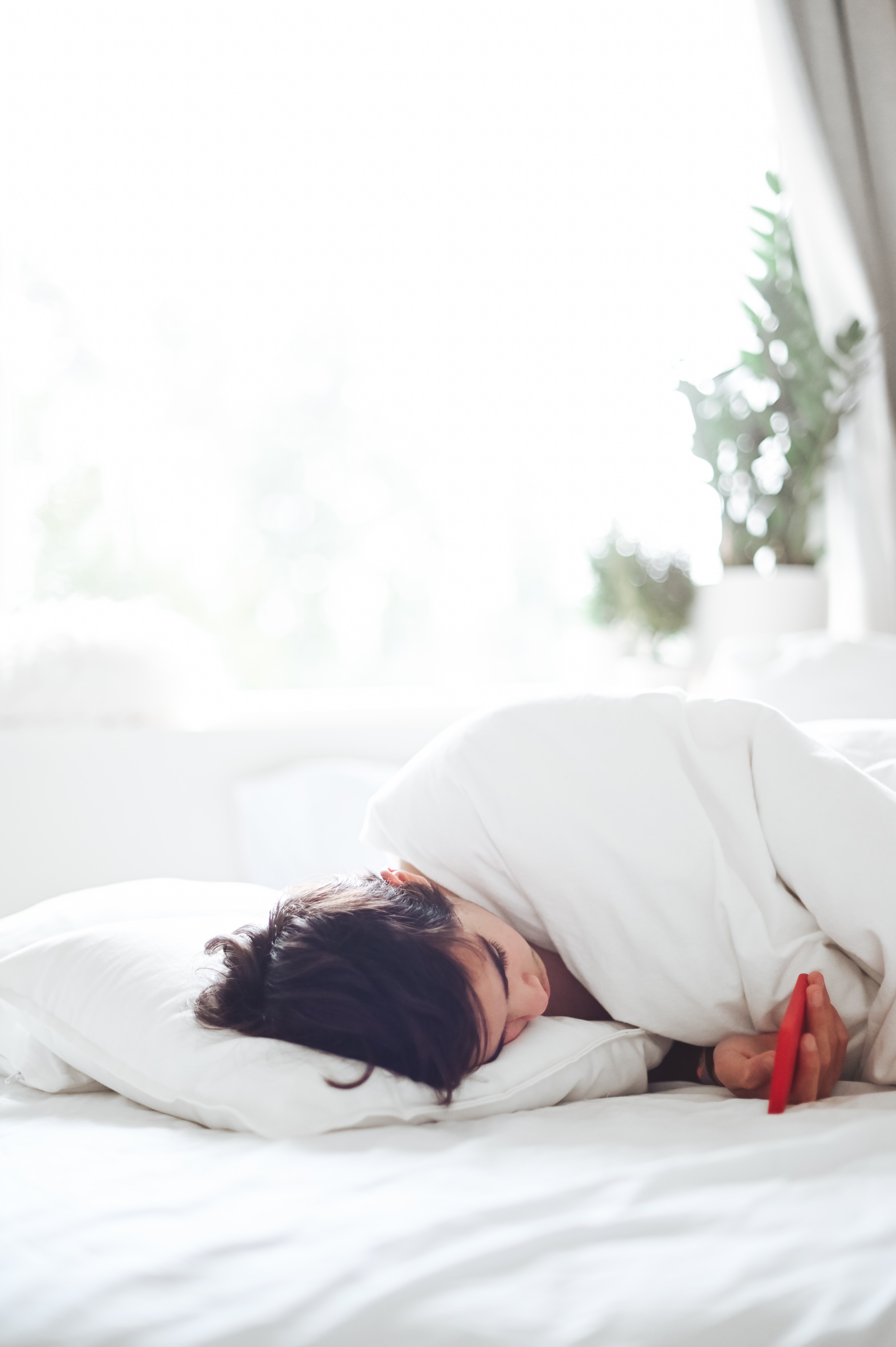 A dark-haired person lays on a bed with white sheets and a white blanket.  The person is laying on their side, holding a red phone, and reading what's on the screen.