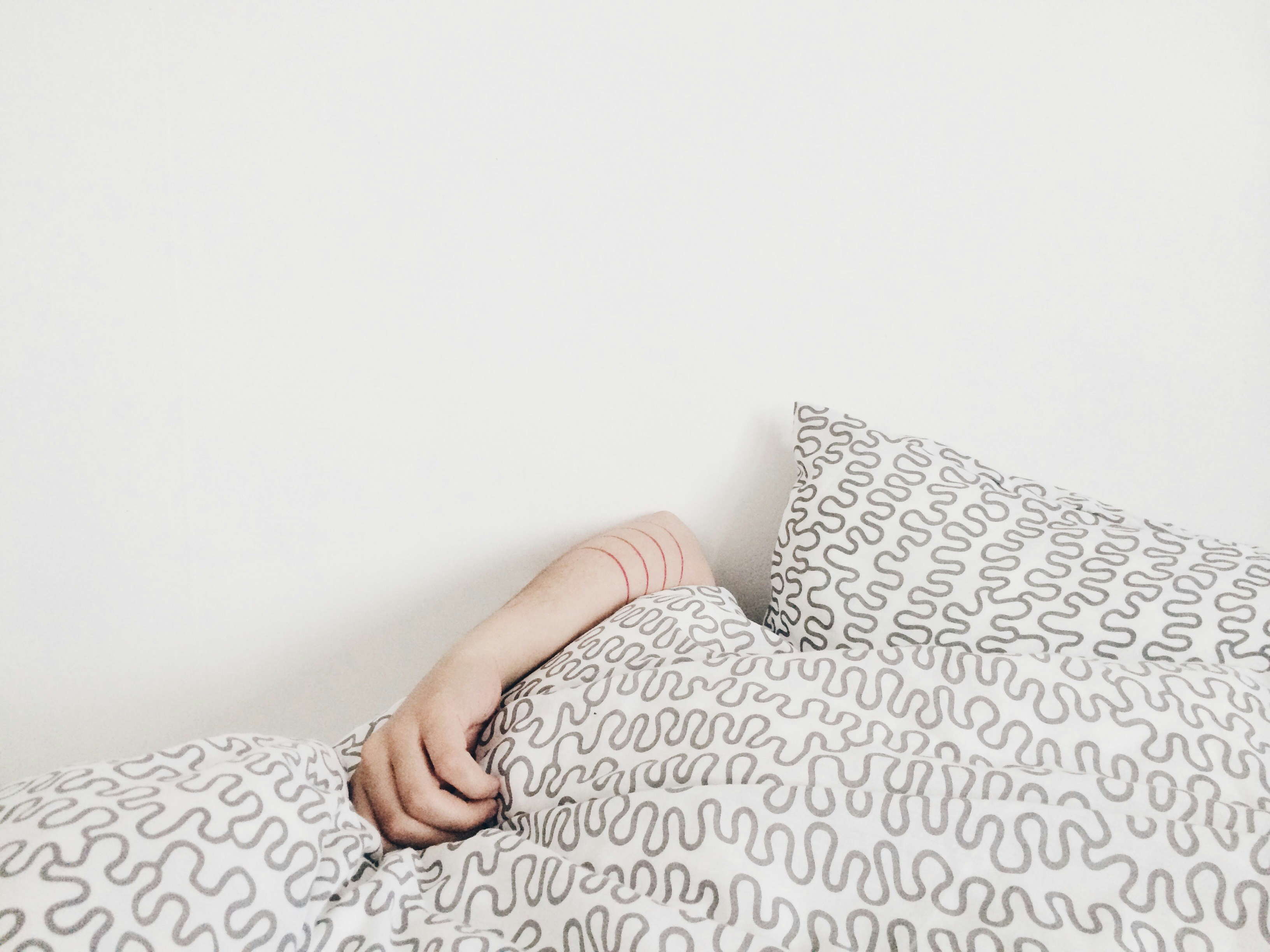 A woman's arm is barely peaking out from underneath a grey and white comforter.  She's laying comfortably in bed, snuggly and warm.