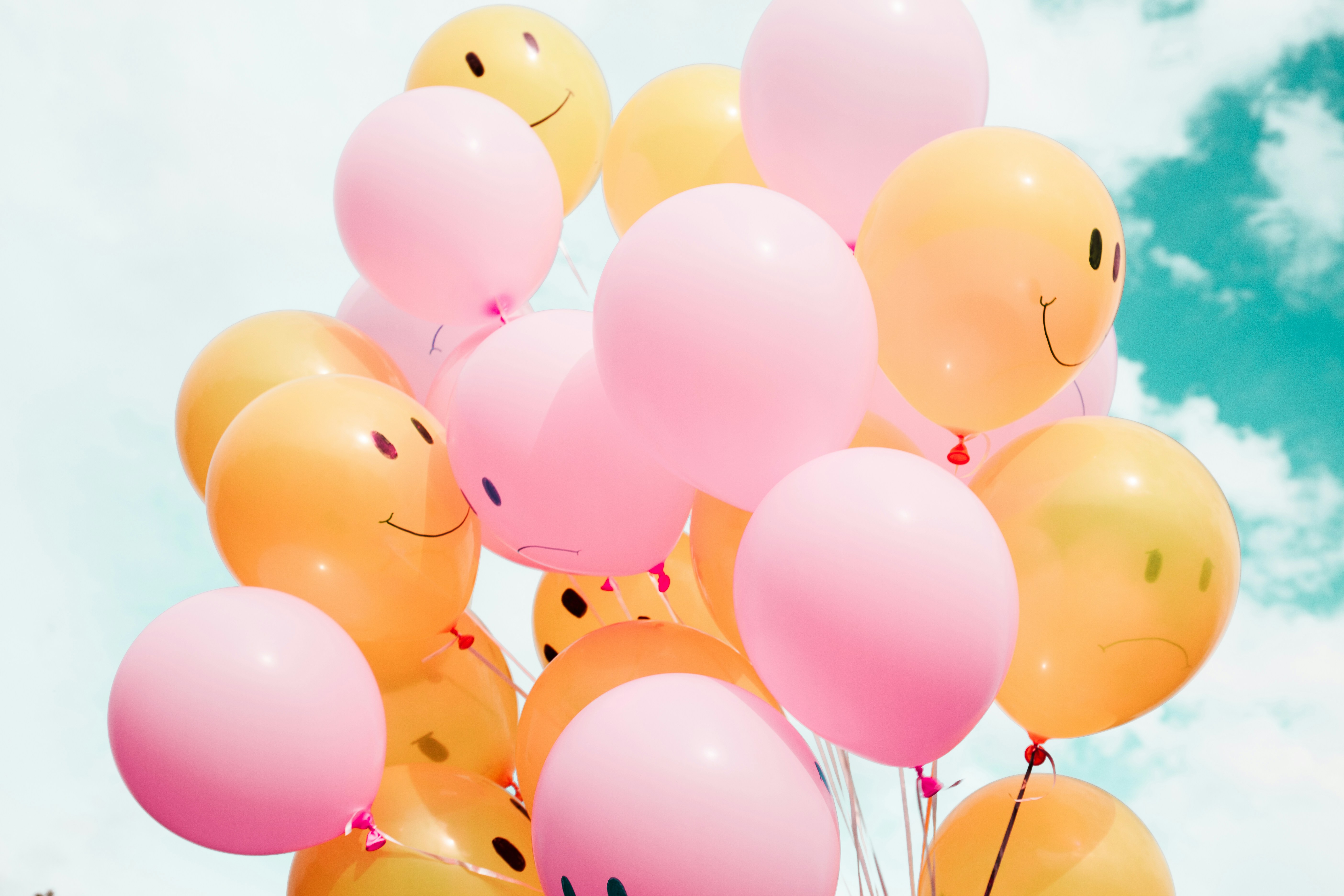 Pink and orange balloons with happy faces printed on them are floating in the air, the blue sky and some white clouds are in the background.