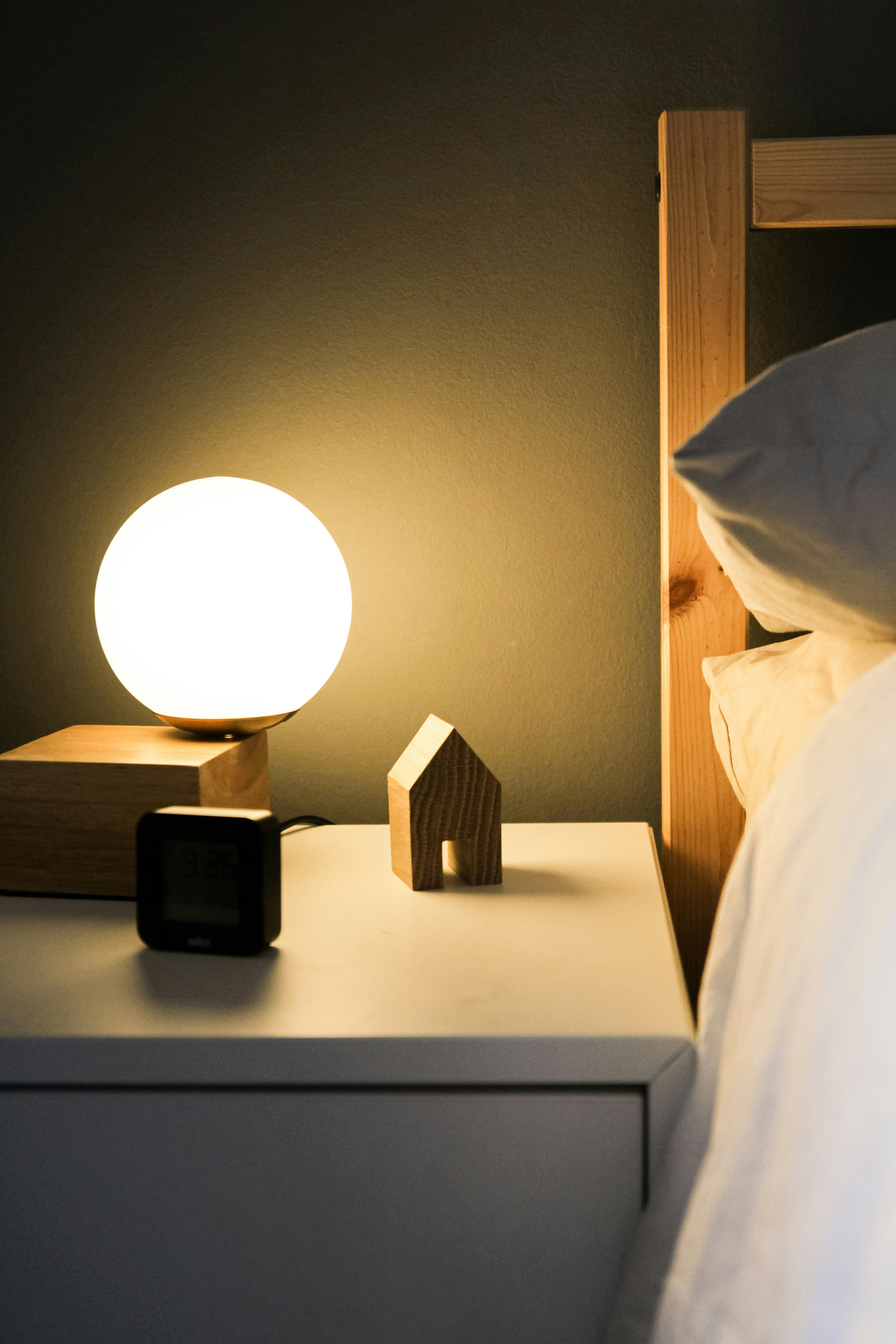 A dimly lit bedroom is softly lit up by a white, round lamp sitting on a short nightstand.  A bed is next to the nightstand.