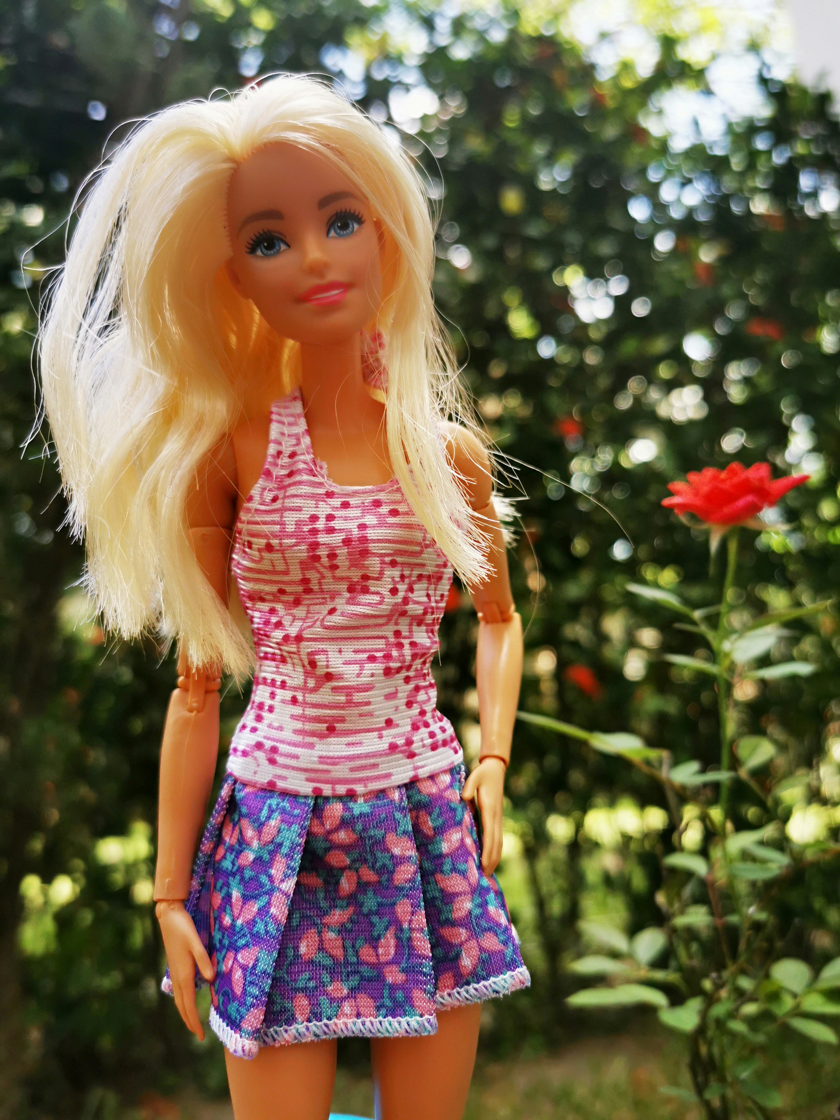 A blonde Barbie doll is wearing a tank top and a mini skirt, standing outside near flowers and a bush.