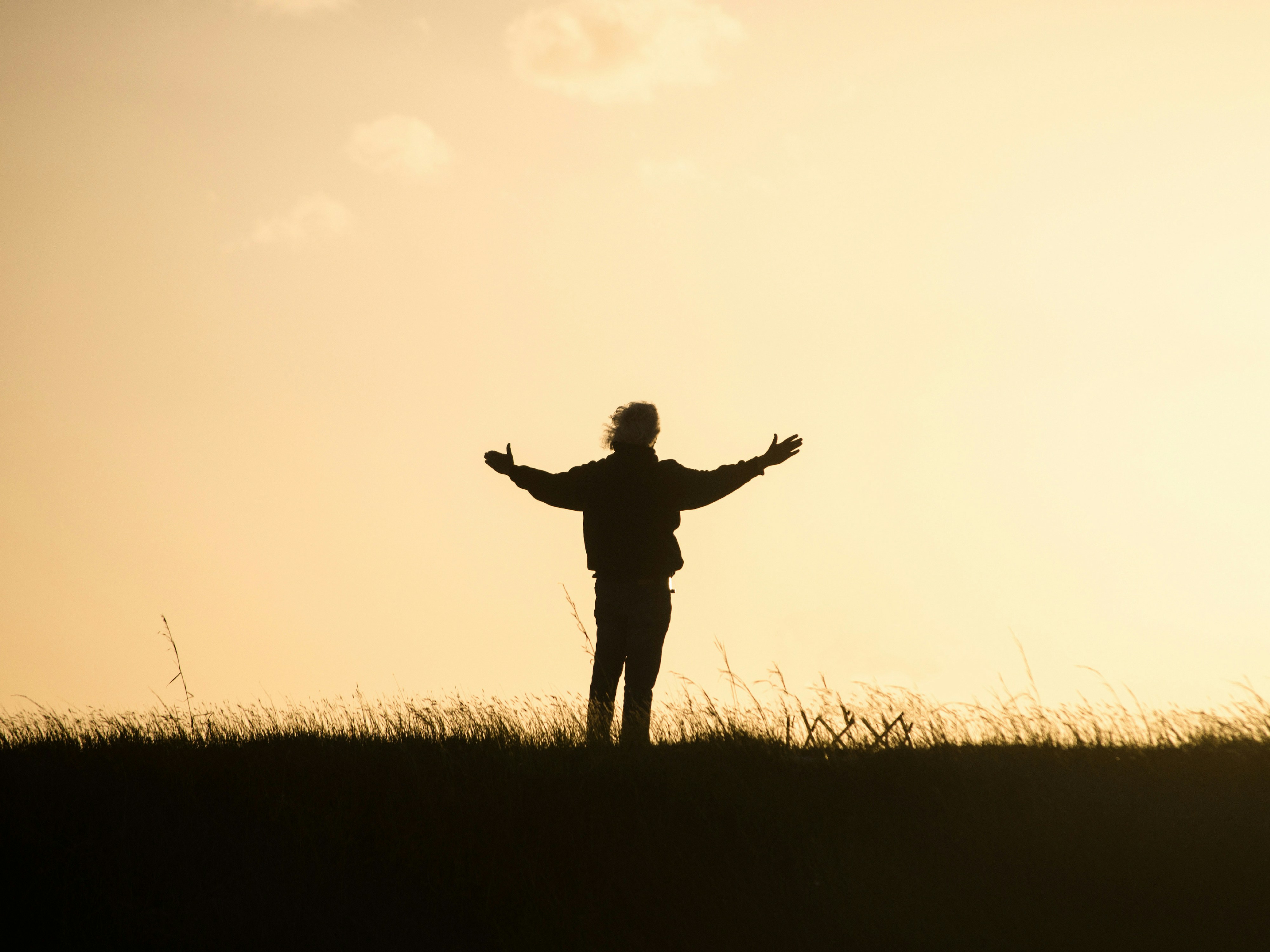 A silhouette of a person shows them standing with their arms open wide, standing in an open field.  The glow of the morning sun shines a muted orange-yellow.