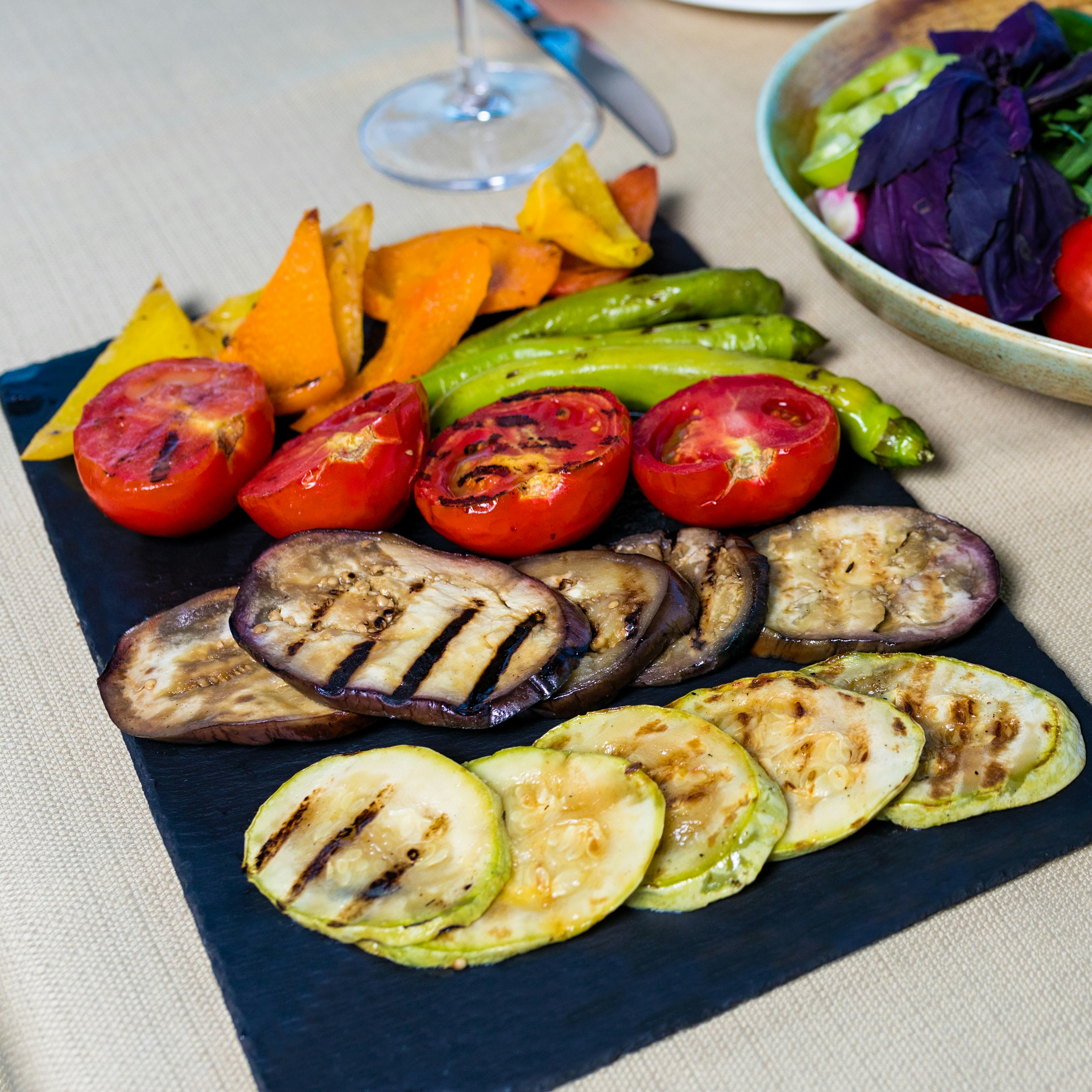 A plate of grilled vegetables is artfully arranged on a dark slate serving board. The assortment includes grilled tomatoes, bell peppers, green chilies, eggplant slices, and zucchini slices. In the background, a salad with vibrant greens and purple leaves is partially visible, along with a wine glass and cutlery on a light-colored tablecloth.