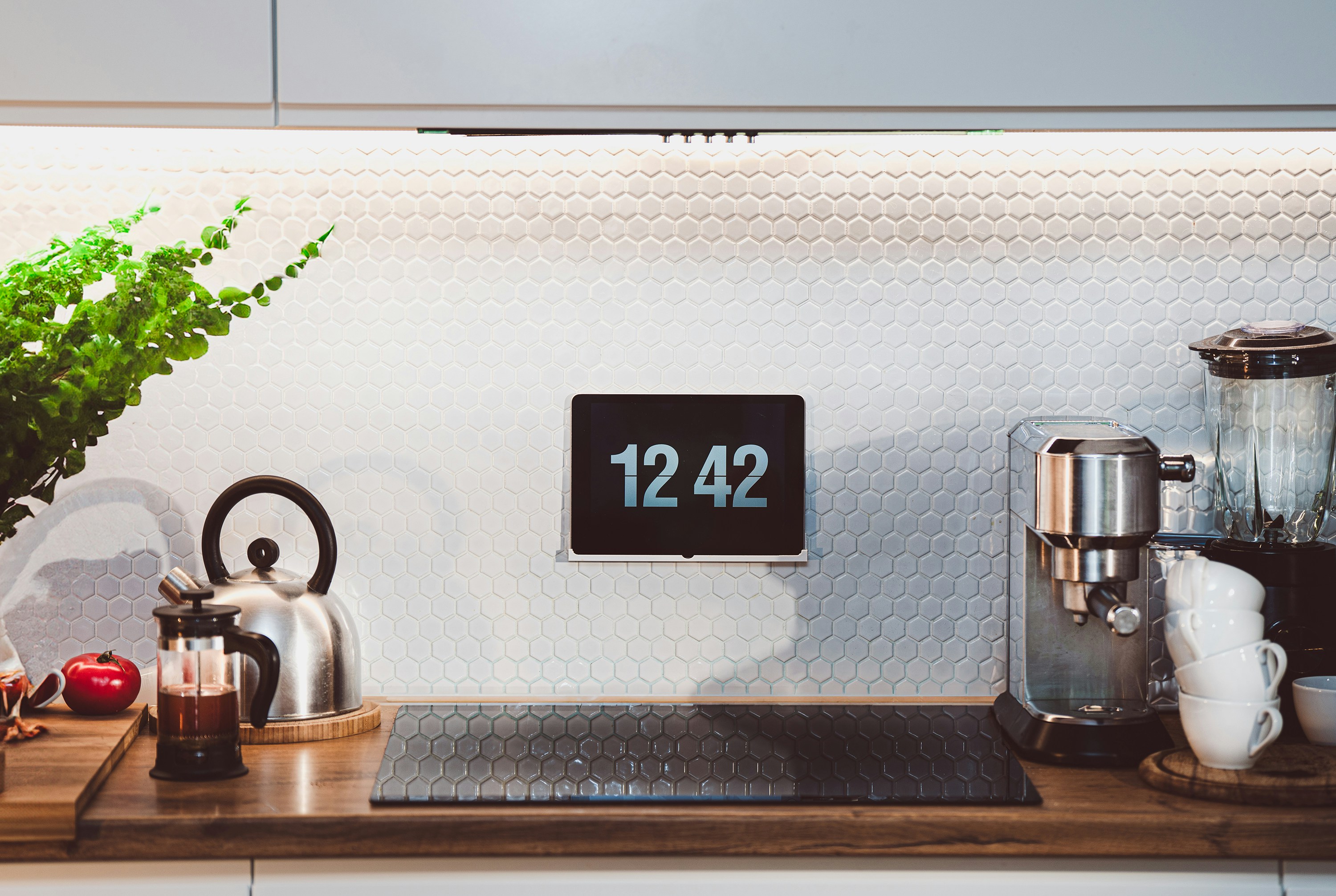 A digital clock is mounted on a wall in a kitchen.  It is placed behind the stove.  To the sides are cooking tools including a blender and a coffee maker.