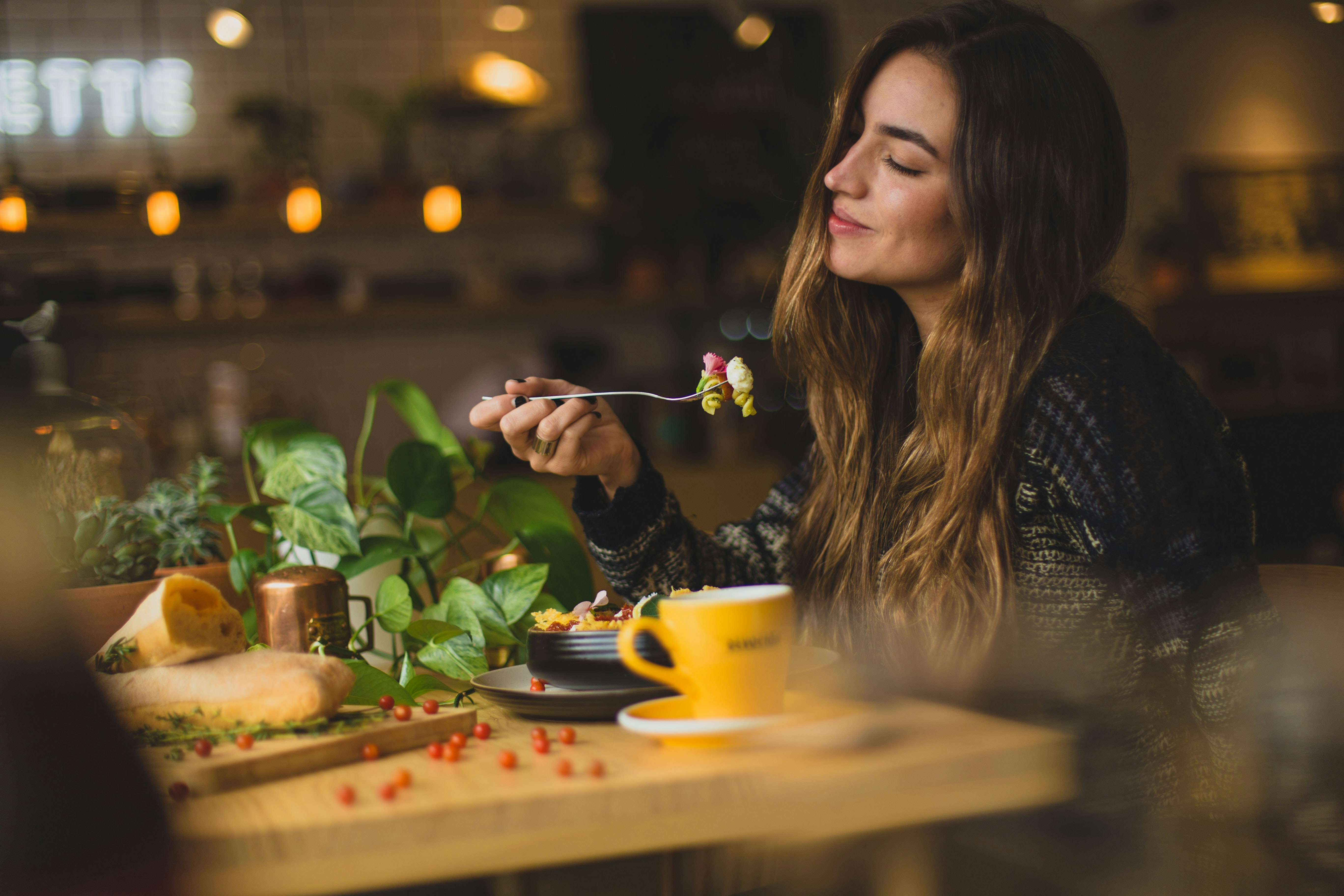 A woman with long brown hair sits at a table, a smile on her face as she's eating.