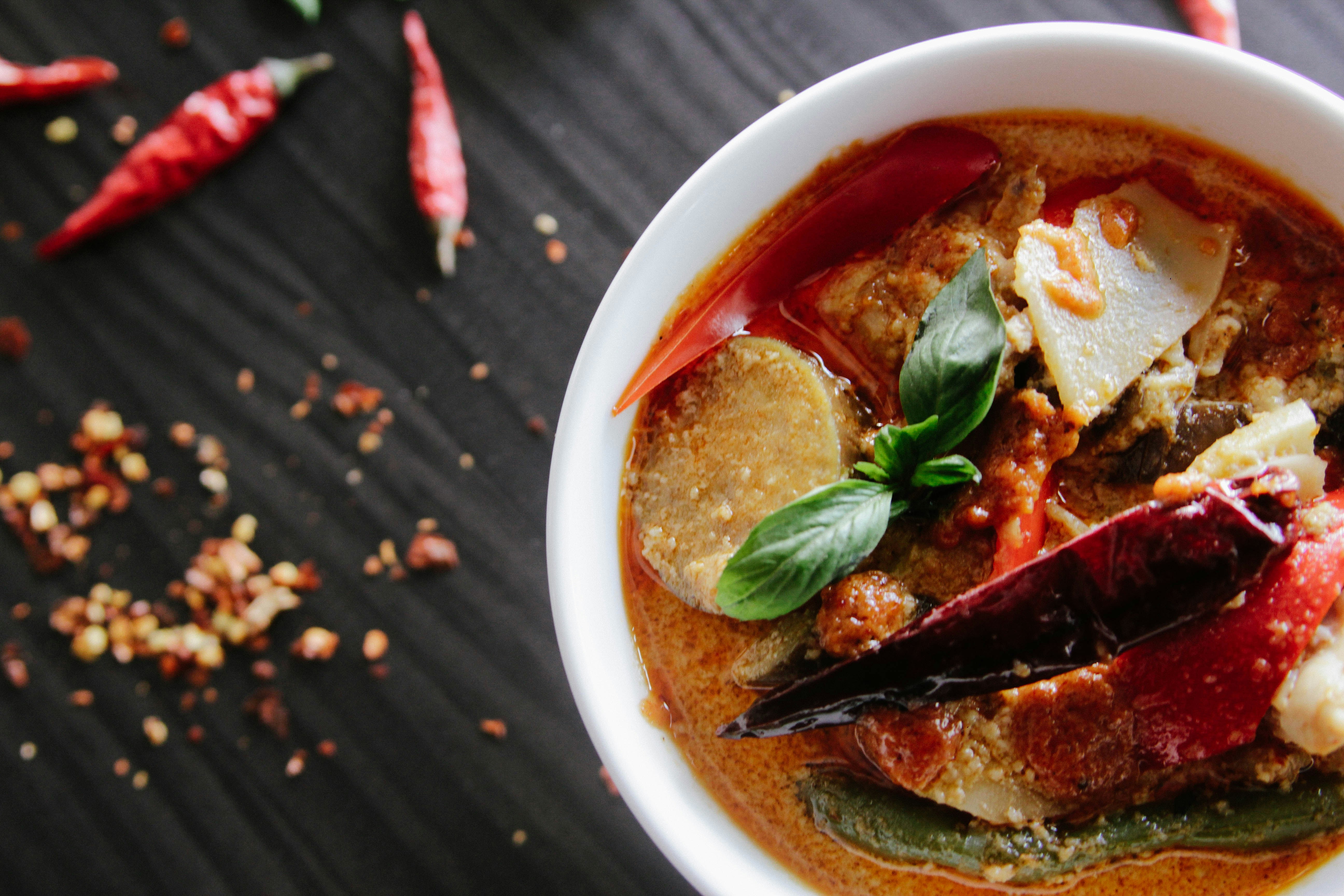 A white bowl of spicy soup is sitting on a dark wooden table.  On the table is a chili pepper with seeds.  The soup is full of spicy vegetables.