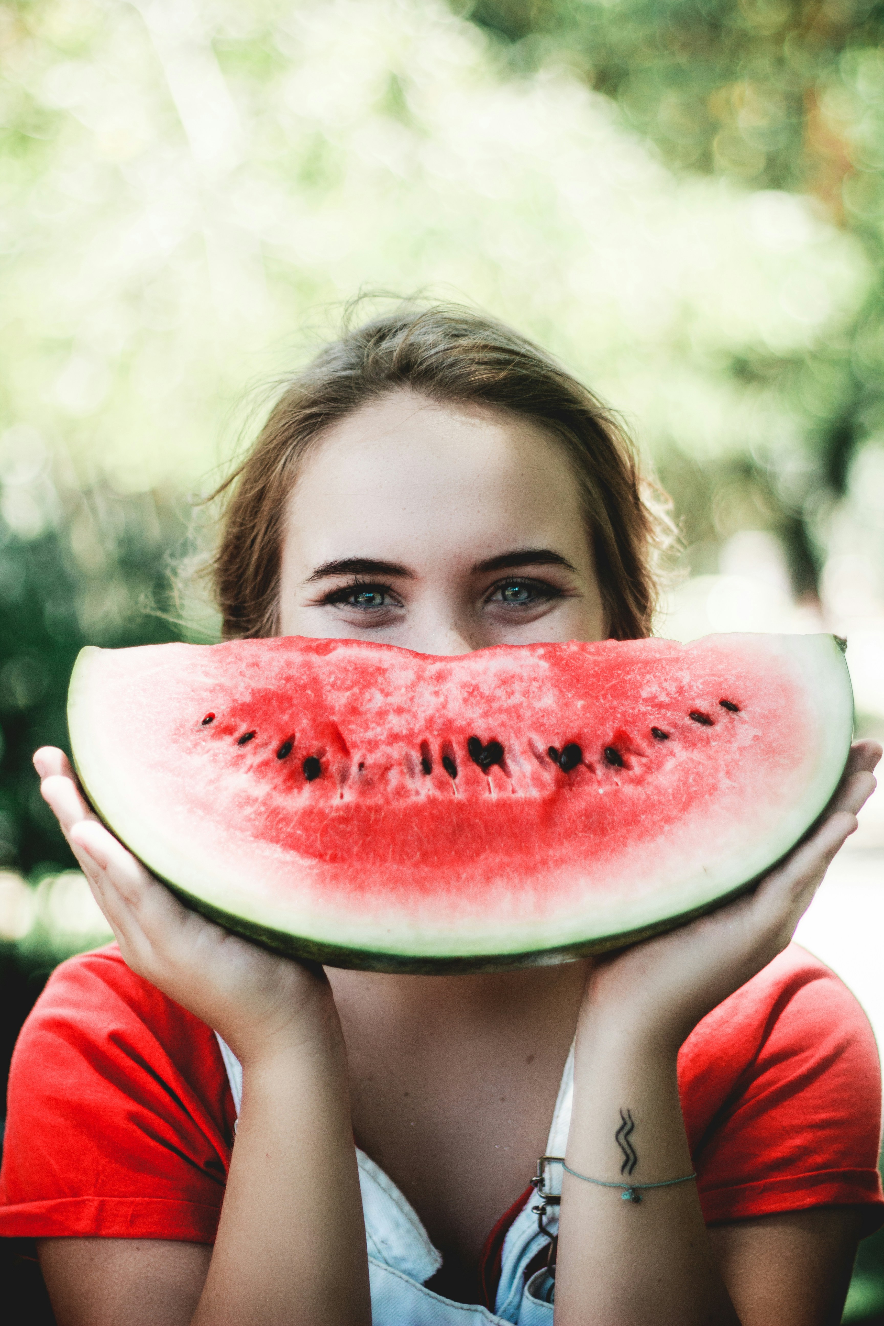 A woman with brown hair wearing a red and white shirt is holding up a big watermelon slice in front of her face.  