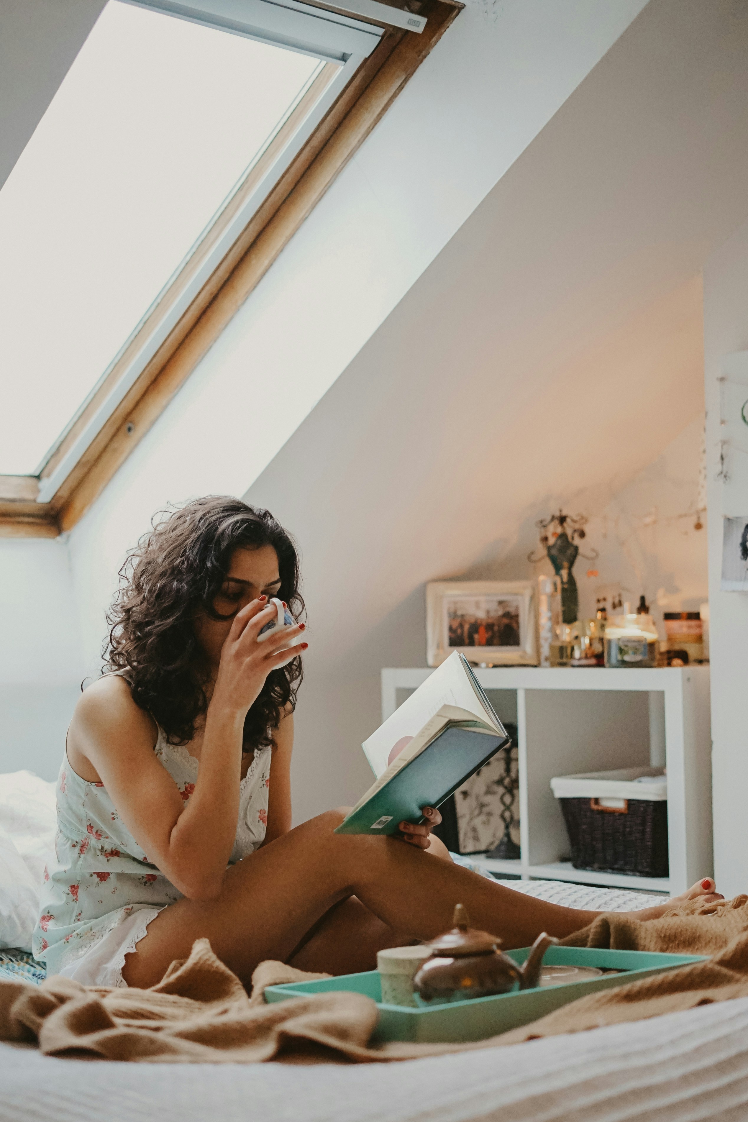 A woman with dark hair sits on her bed, sipping tea.  A tray is nearby with a teapot.  She's holding a book.