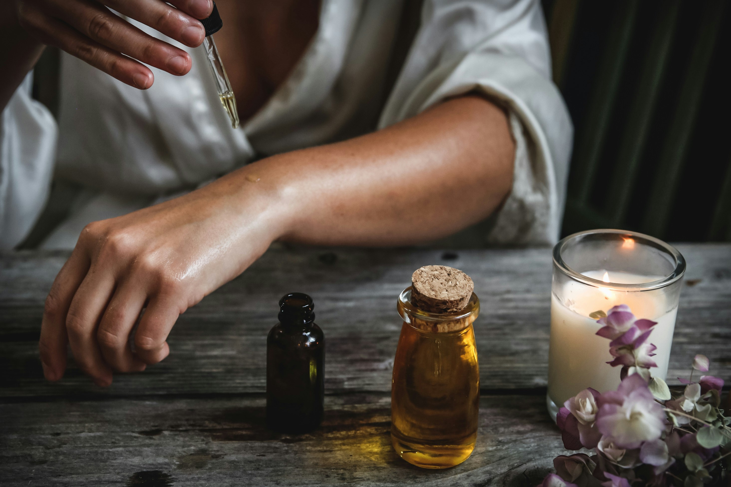 A woman's arm is outstretched.  She's dripping small drops of oil on it.  Nearby is a lit candle and  some flowers sitting on a grey wooden table.