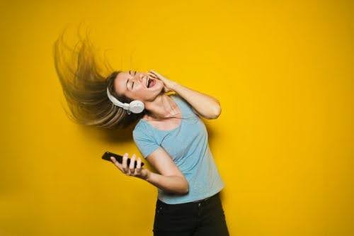 A blonde woman is dancing.  She has headphones on, is wearing a blue shirt and black pants, and a yellow wall is behind her.