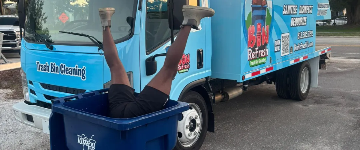 Person playfully inside a blue trash bin in front of cleaning service truck Person playfully inside a blue trash bin in front of cleaning service truck
