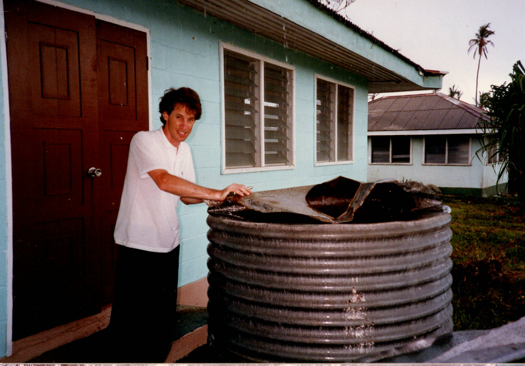 A person stands next to a large, round metal water tank with a damaged cover outside a light blue house with brown double doors and several windows. Another house and palm trees are visible in the background.