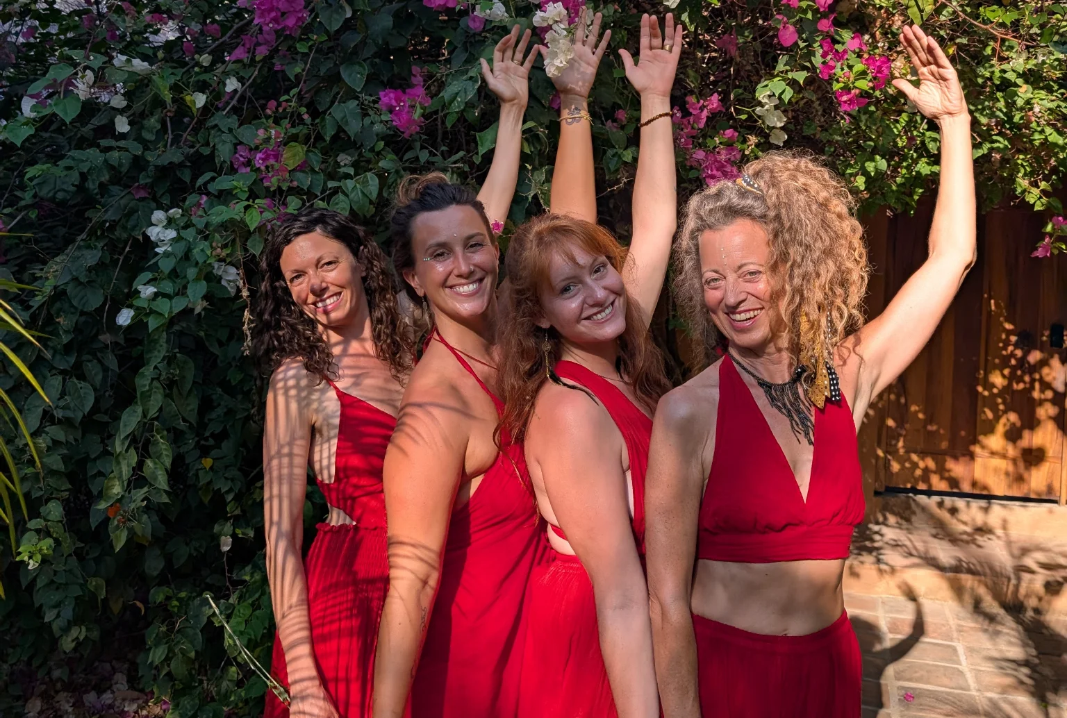Four joyful women in red dresses standing under vibrant bougainvillea flowers, celebrating sisterhood and feminine connection at a Tantra retreat.