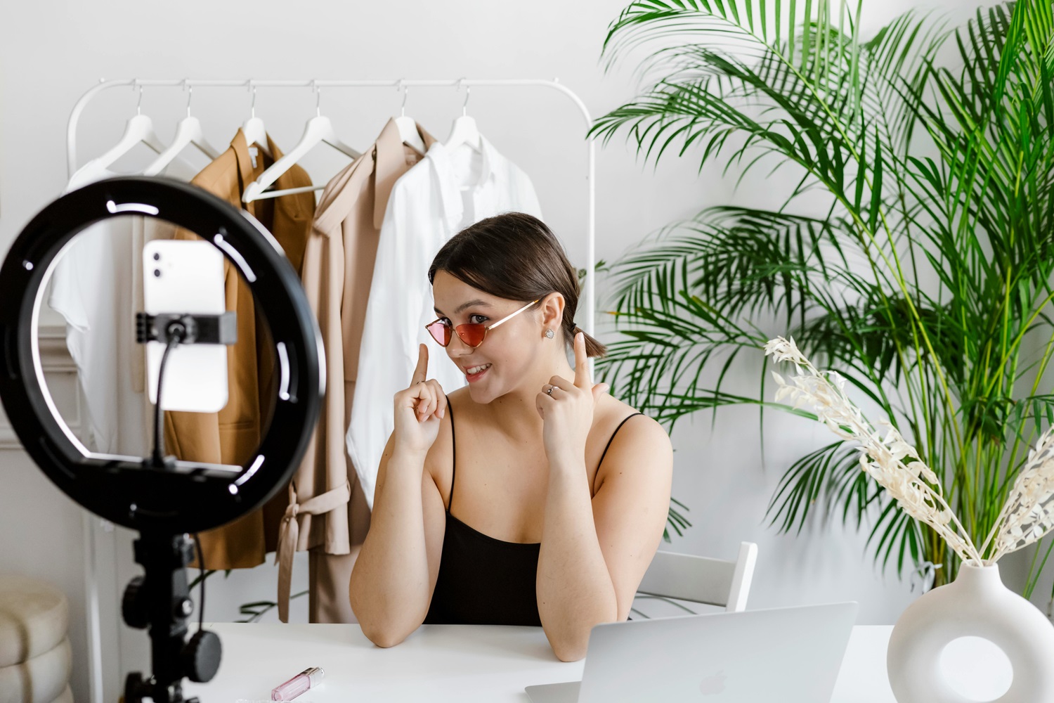 Female influencer recording a fashion video with a smartphone and ring light, featuring a clothing rack with neutral-colored outfits in the background.