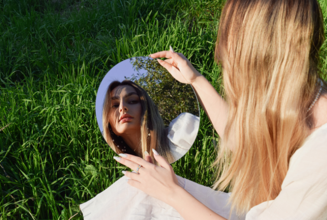 Woman looking into a mirror that reflects a glowing universe of hearts and light, soft ethereal atmosphere”