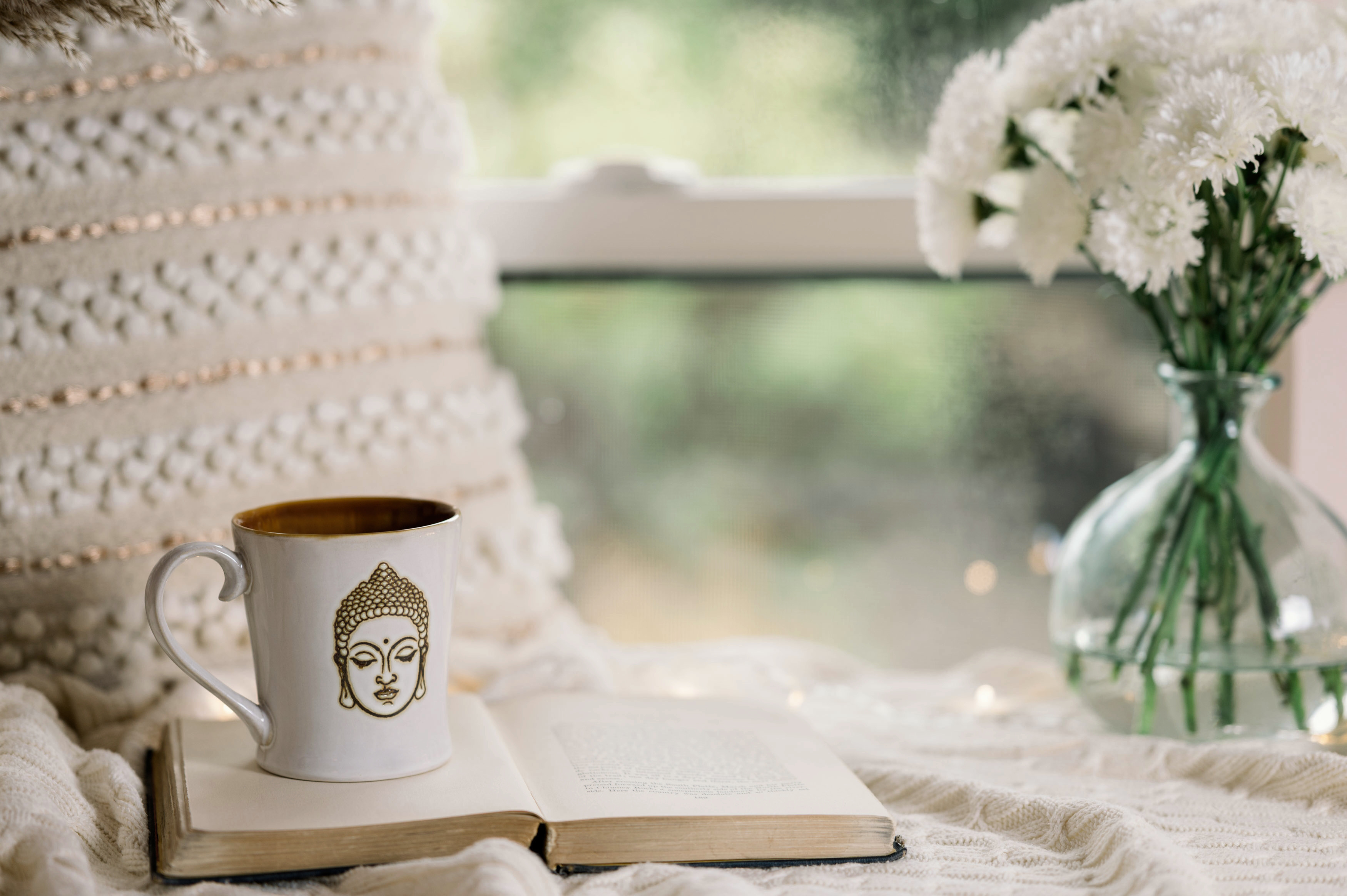 A serene setting featuring a cozy nook with a white Buddha mug filled with tea or coffee placed on an open book. In the background, there are white flowers in a clear glass vase and a window letting in soft, natural light. The overall ambiance is calm and inviting, perfect for reflective moments or morning routines.