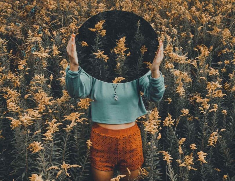 Woman holding up a mirror to her face and hiding behind it. She is in a field wearing colorful clothing