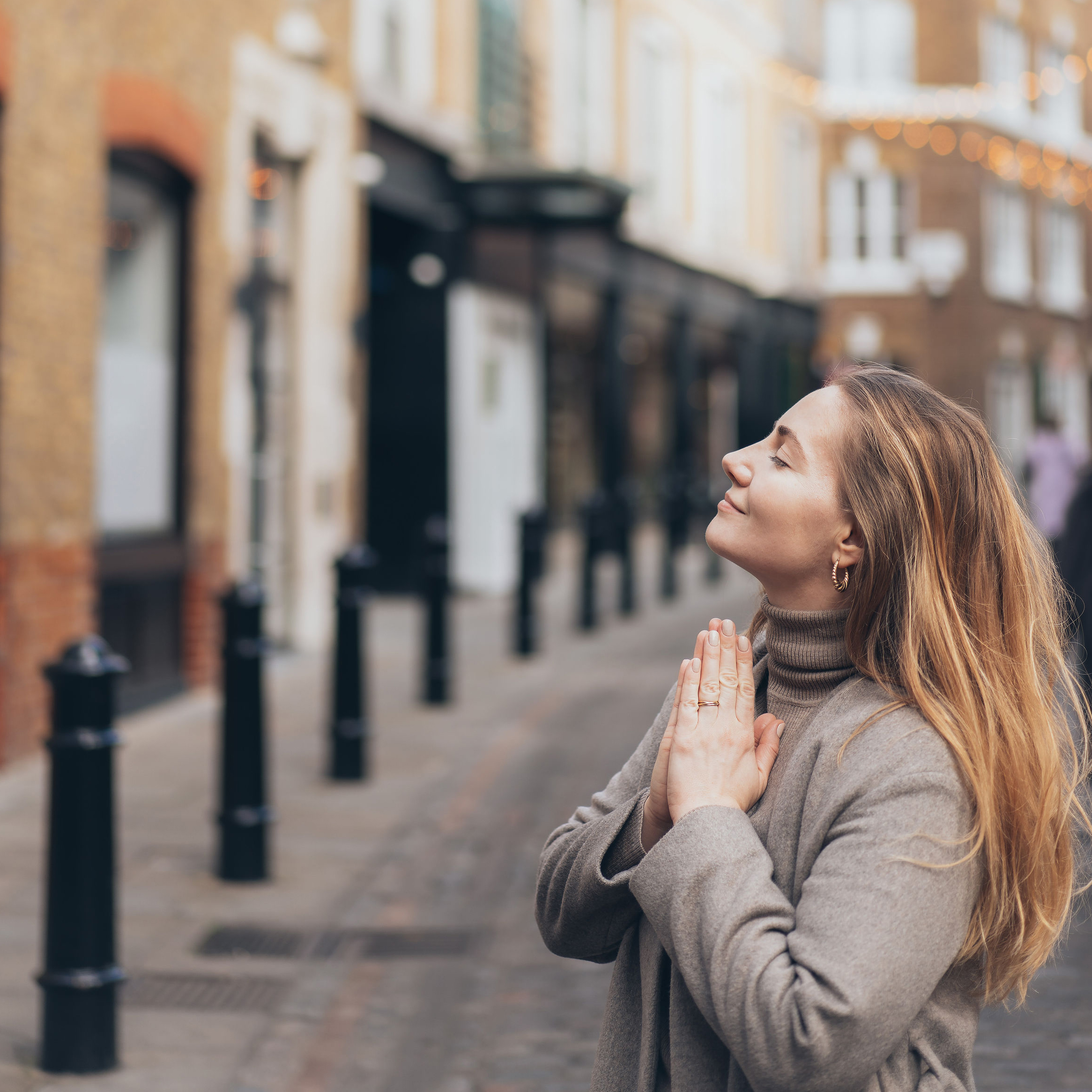 A woman with long blonde hair stands on a cobblestone street in a city, wearing a grey coat and turtleneck. She has her eyes closed and hands pressed together in a prayer-like gesture, appearing serene and content. The background features blurred buildings and streetlights.