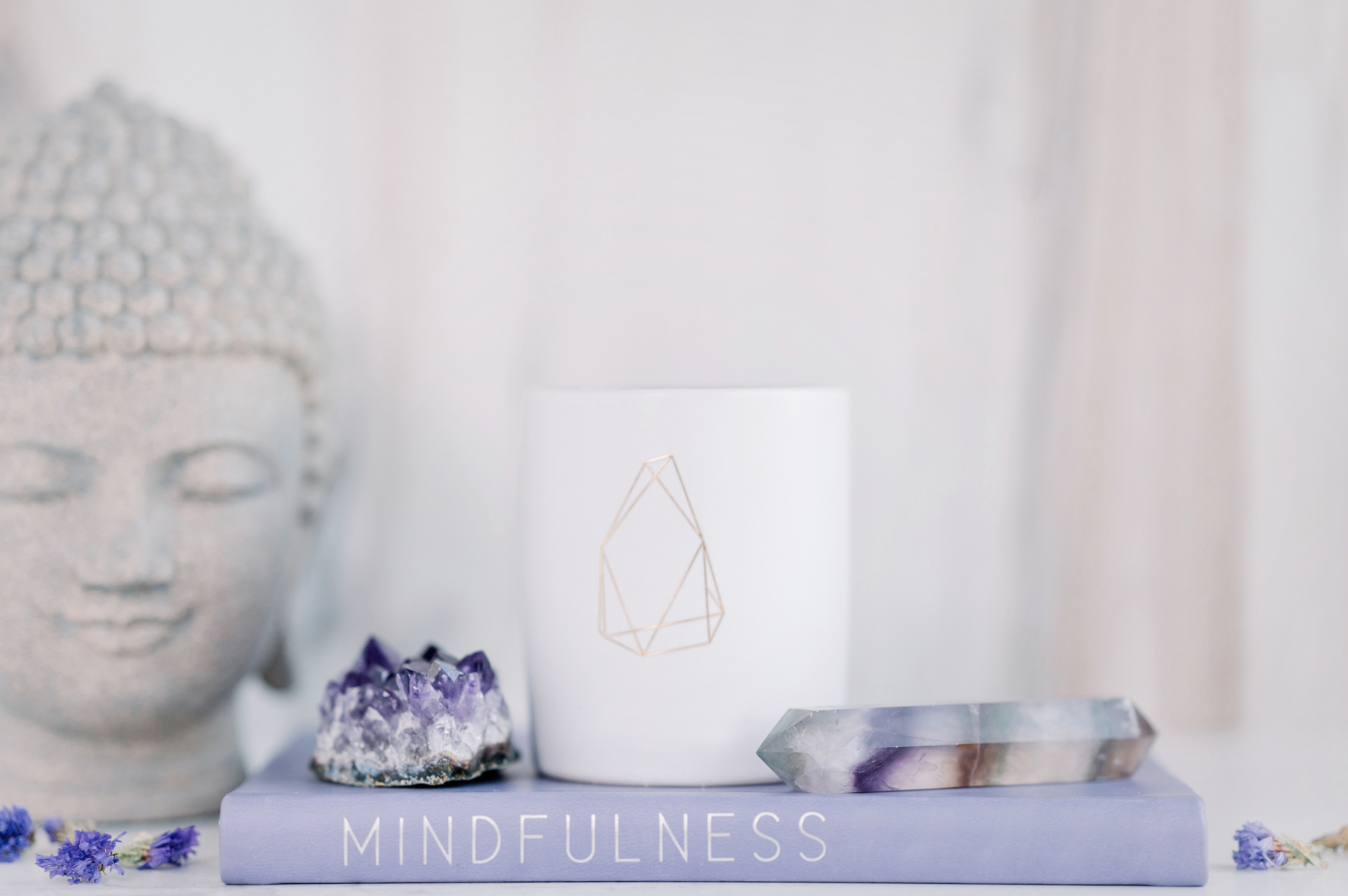 A serene setup featuring a Buddha statue, an amethyst crystal, a white candle with geometric design, and a book titled 'Mindfulness' with a crystal wand on top.